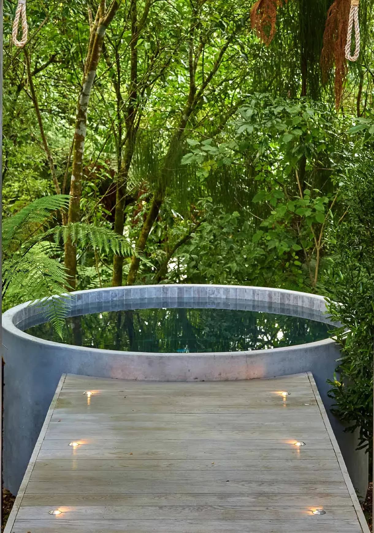 Circular hot tub surrounded by greenery, with wooden decking and recessed lighting along the edge.