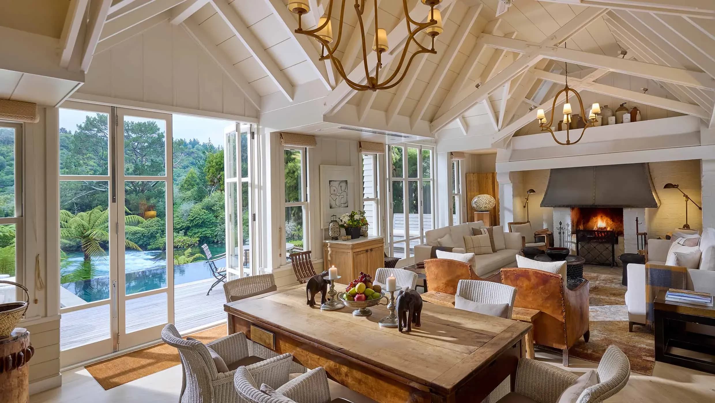 Dining area with wooden table, armchairs, and large windows overlooking greenery and a pool.
