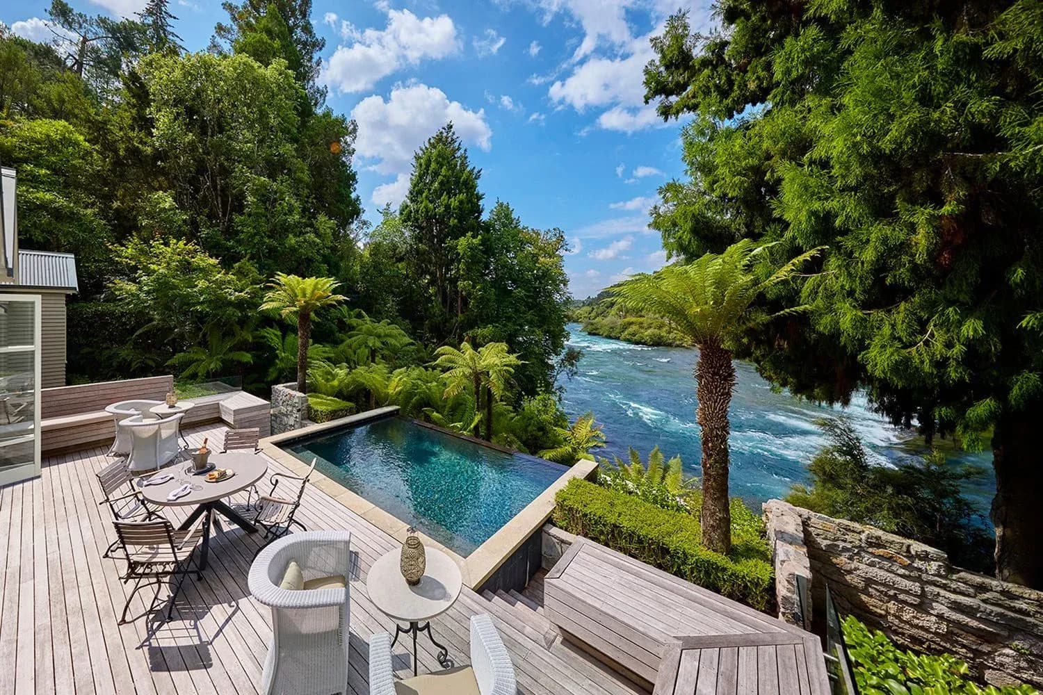 Deck area with seating and dining table overlooking a swimming pool and a river surrounded by lush greenery.