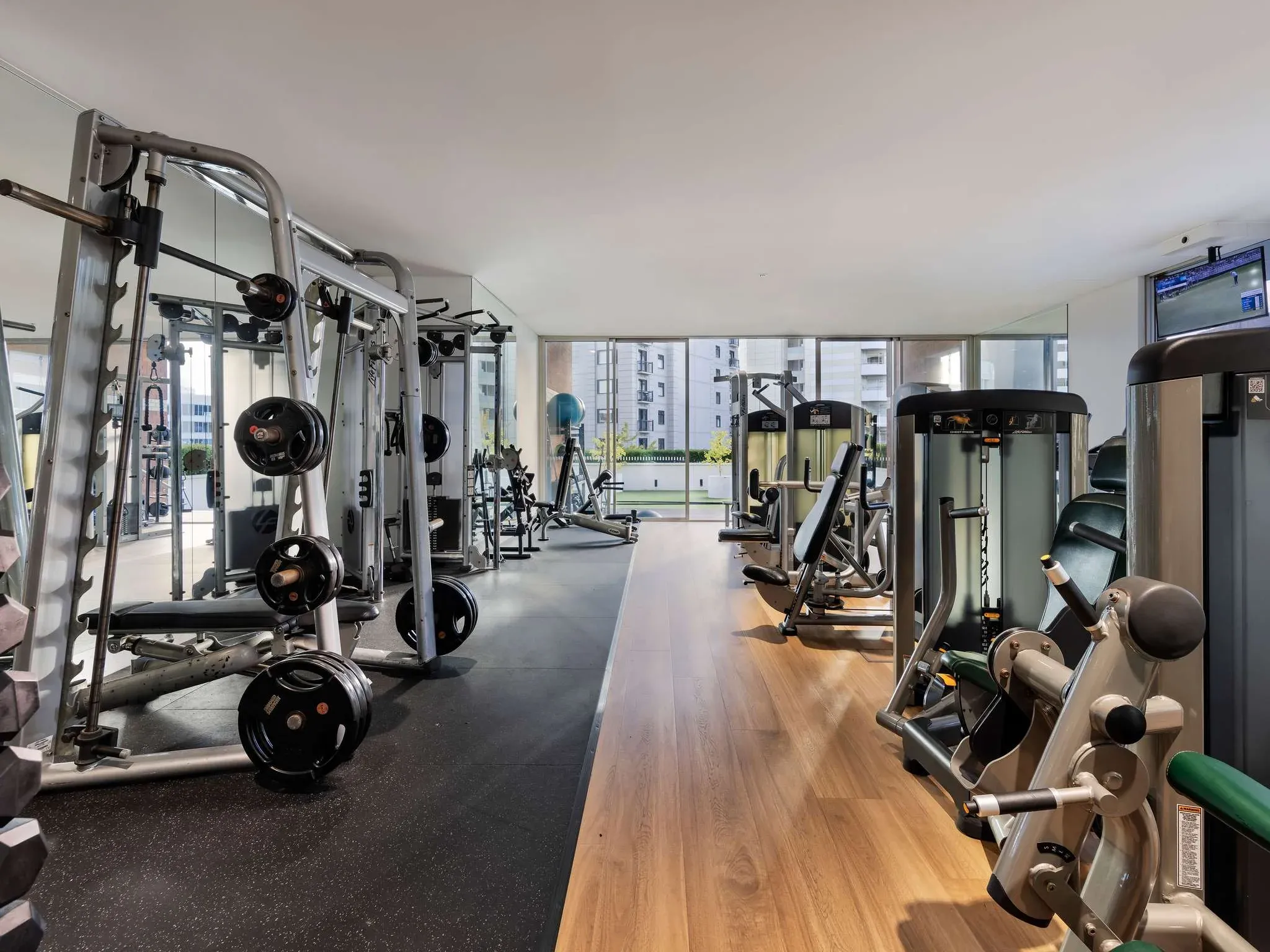 Gym interior with various weightlifting machines, free weights, and large windows allowing natural light.