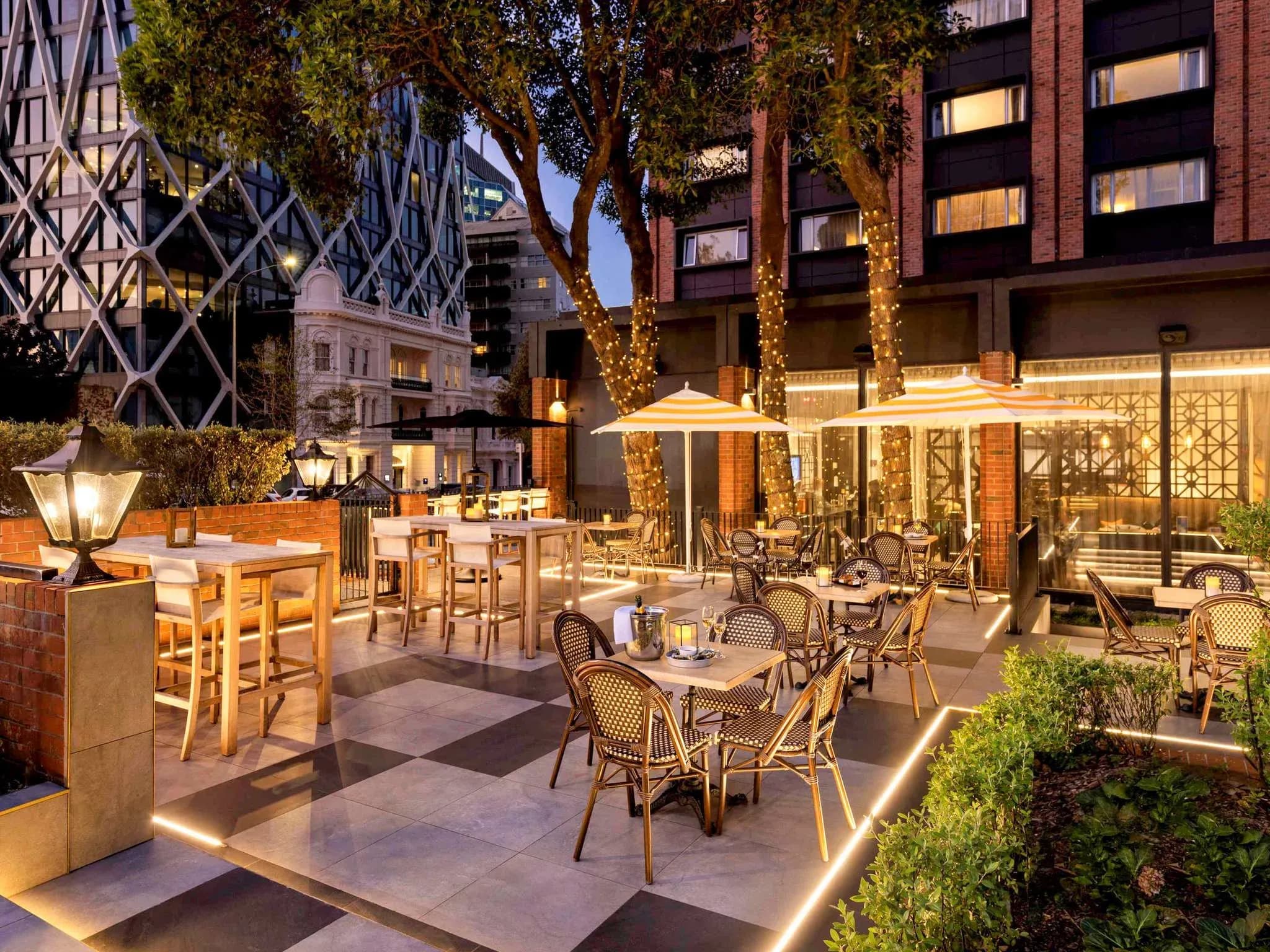 Outdoor dining area with wooden tables, wicker chairs, and parasols, surrounded by greenery and illuminated by soft lighting.