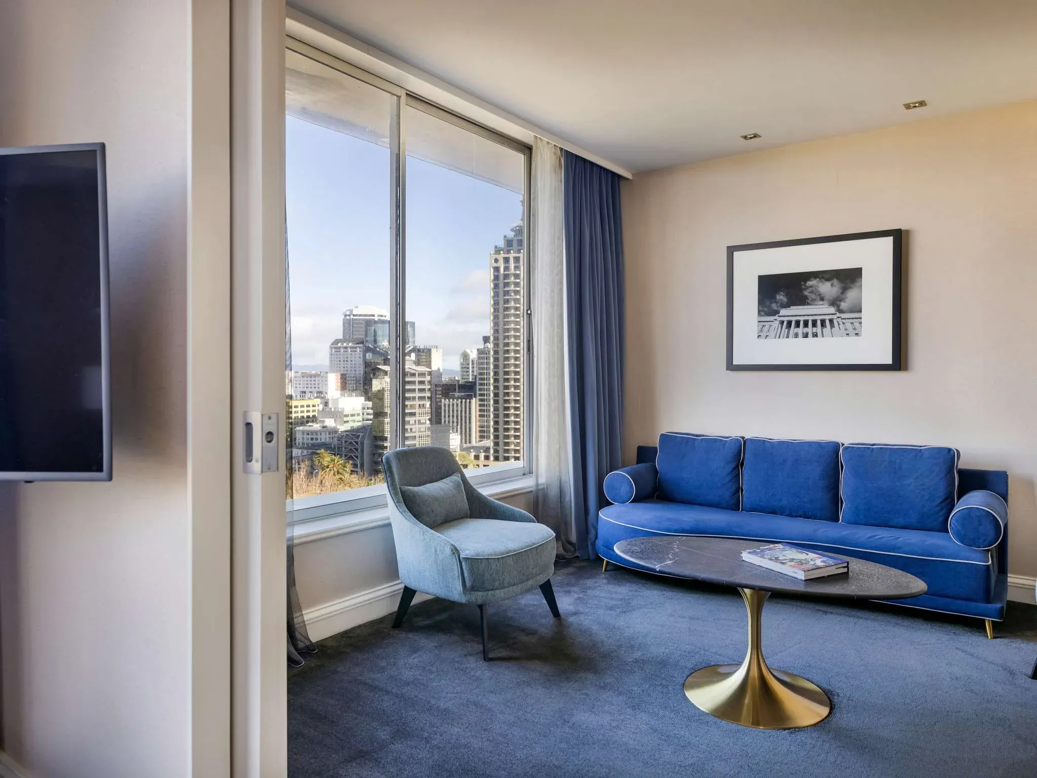 Sitting area with a blue sofa, grey armchair, round marble coffee table, and large windows offering city views.