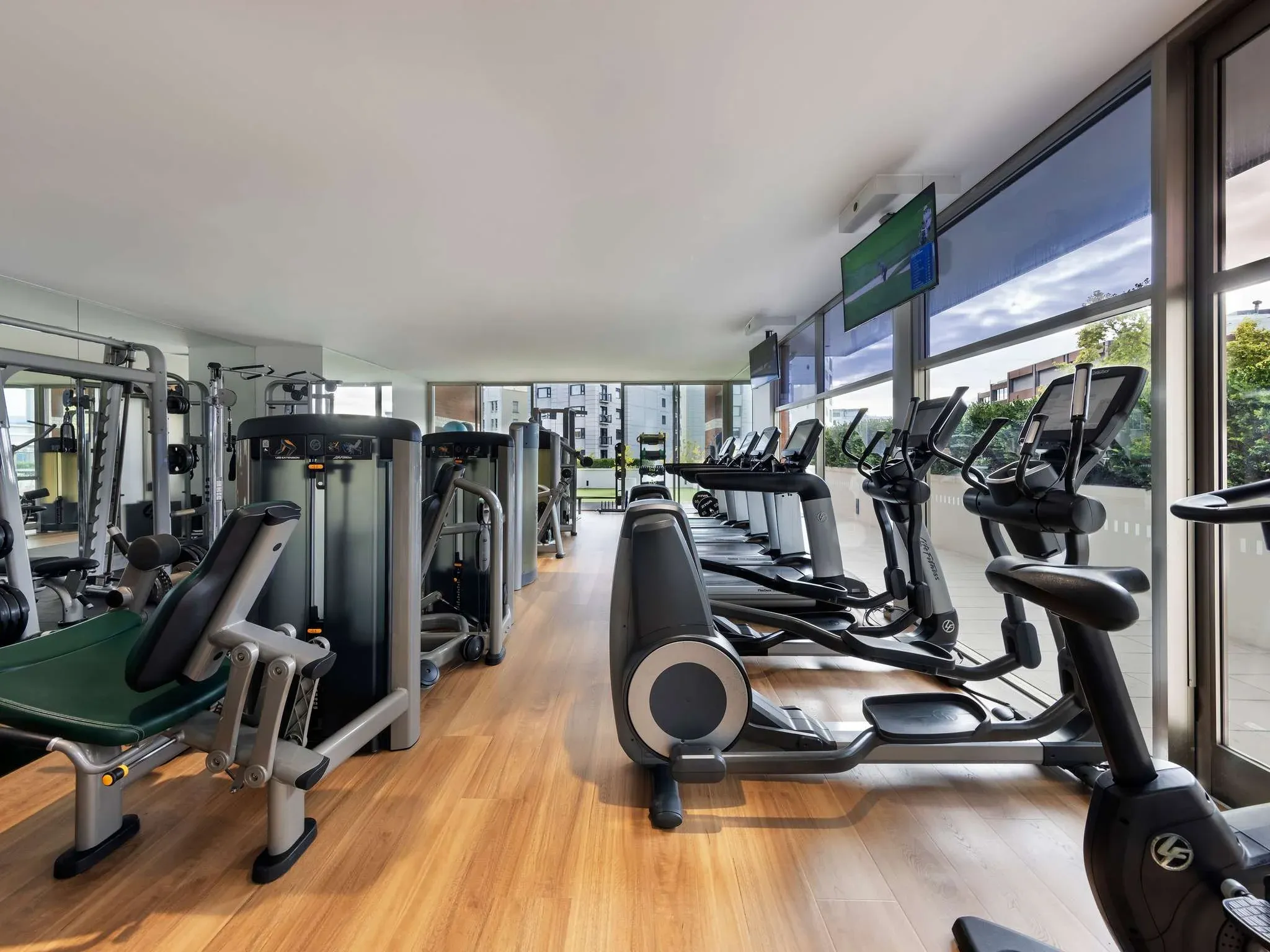 Gym interior with various exercise machines, large windows providing natural light and views of greenery outside.