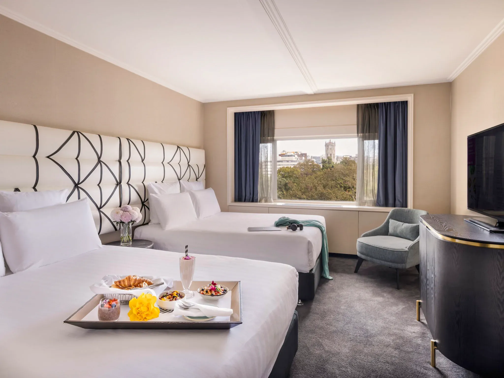 A guest room with two beds, a tray of food, a window view and a TV, featuring neutral tones and modern decor.