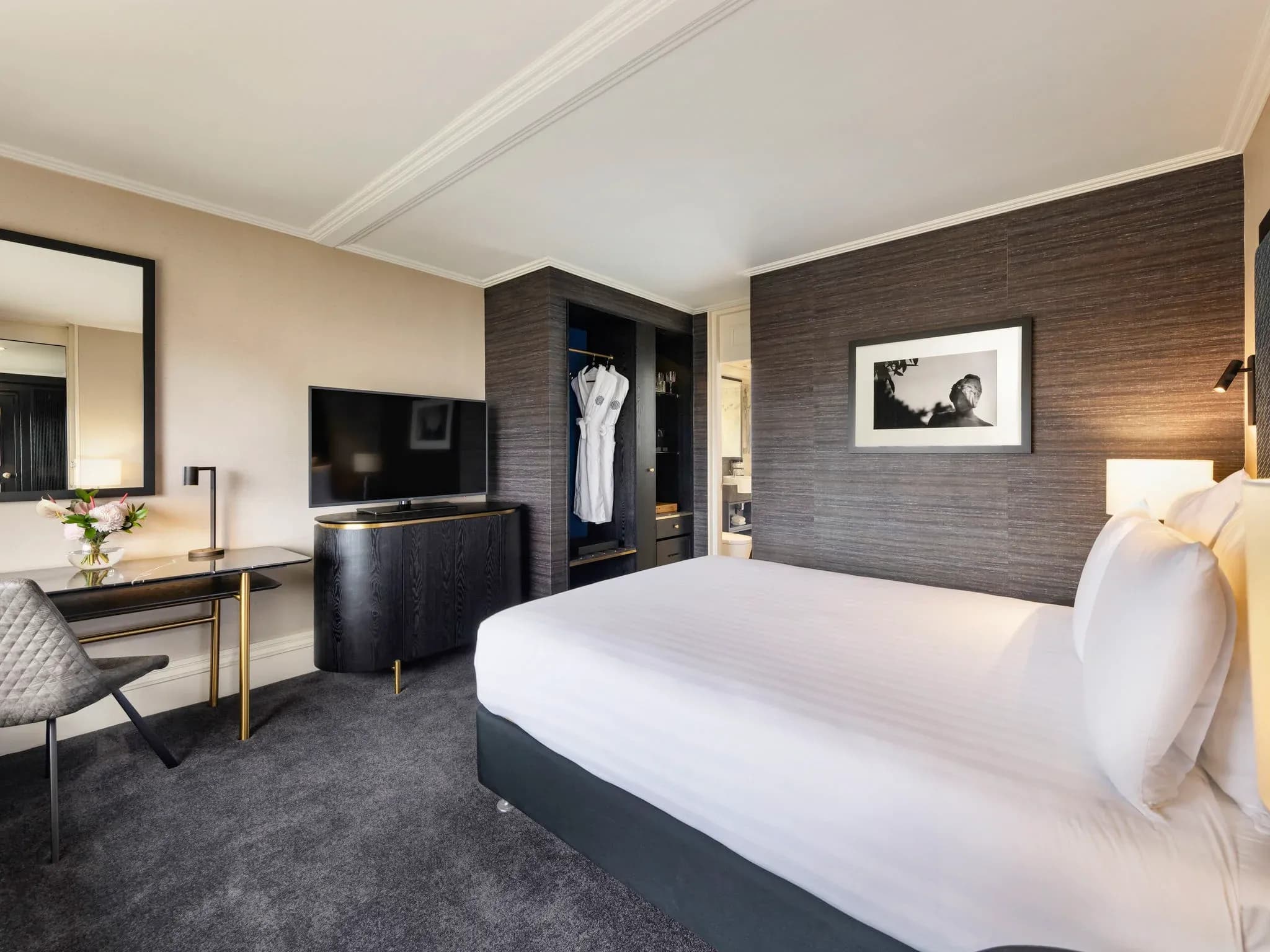Guest room with a king bed, desk, television, and a mirror, featuring neutral tones and modern furnishings.