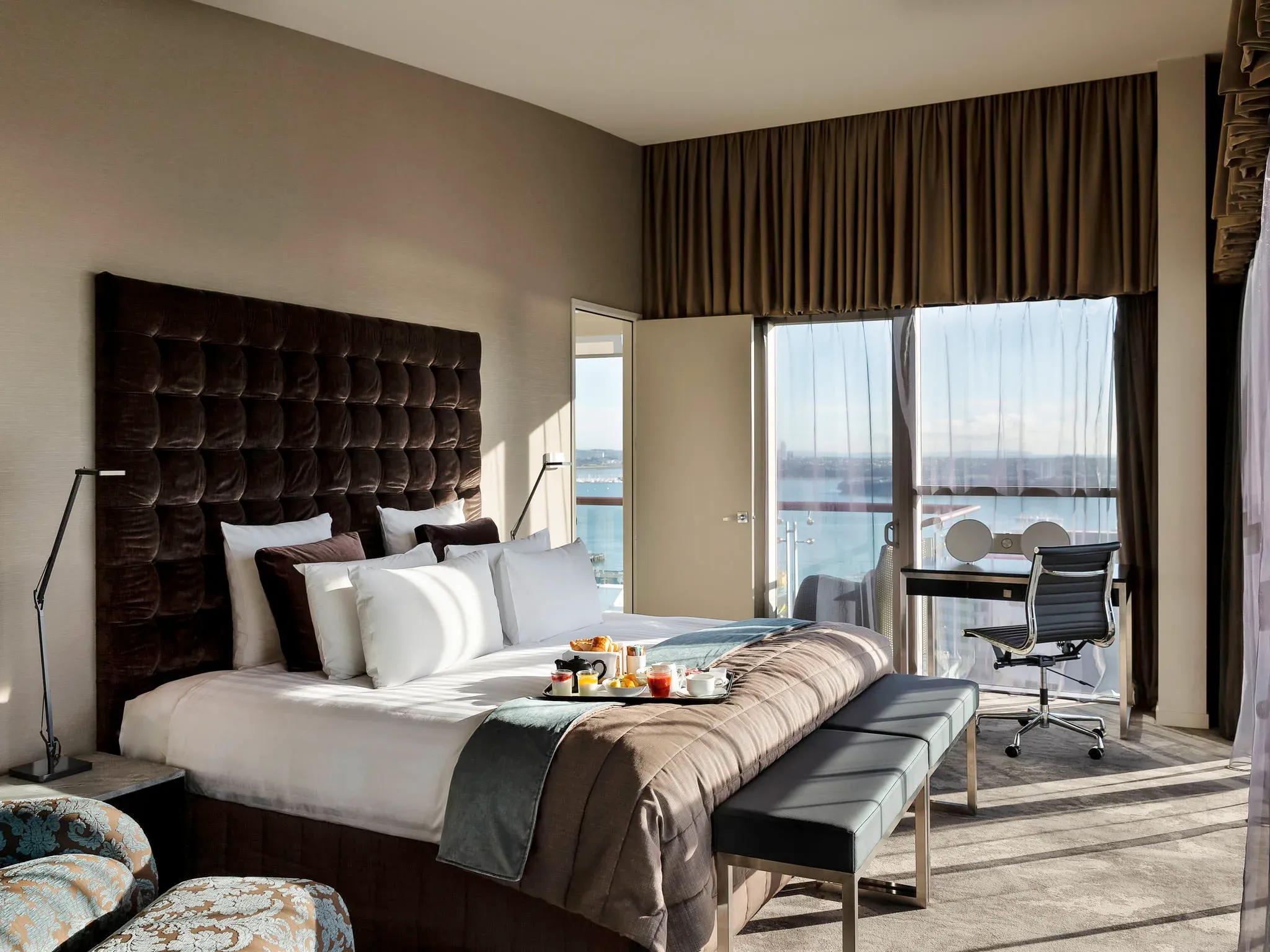 Bedroom with a tufted dark headboard, white bedding, a grey throw, and a balcony view of the water.