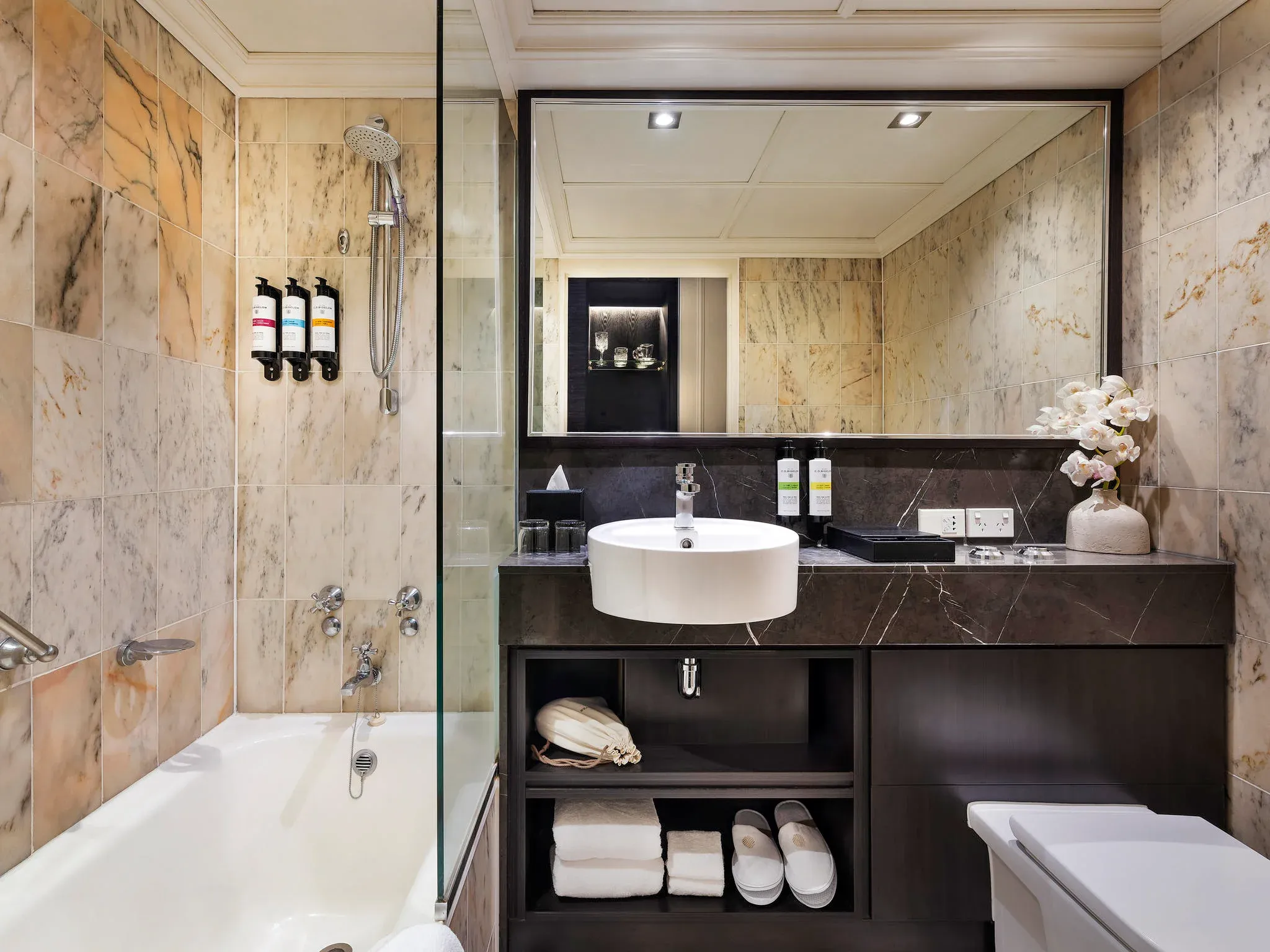 Bathroom with marble walls, a glass shower, a freestanding sink, and a bathtub with toiletries and towels.