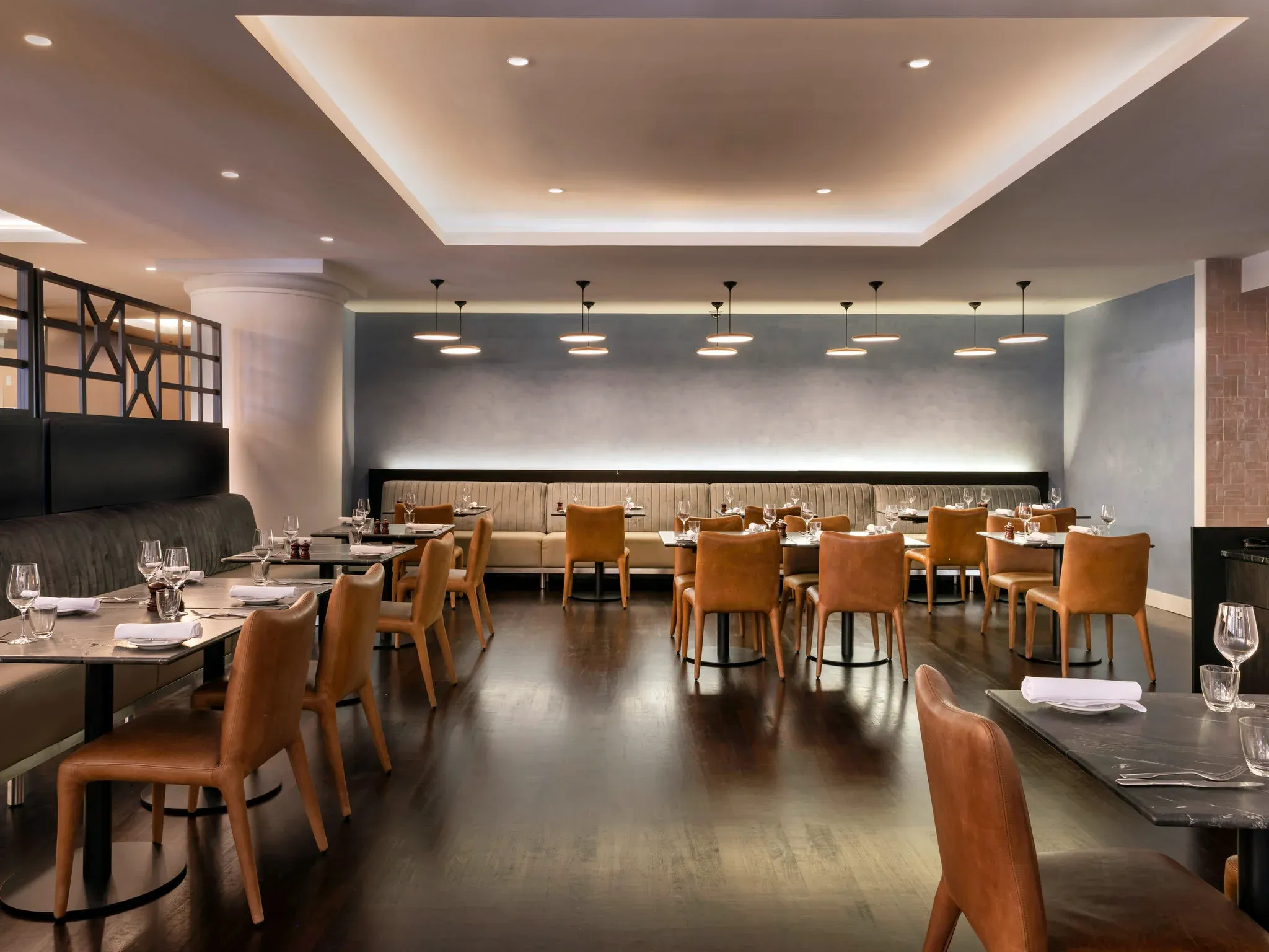 Restaurant interior with wooden flooring, grey walls, leather chairs, and tables set with glassware.