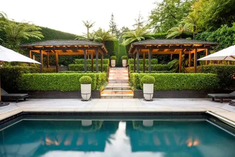 Poolside view with two covered seating areas, lush hedges, and steps leading to a landscaped garden.