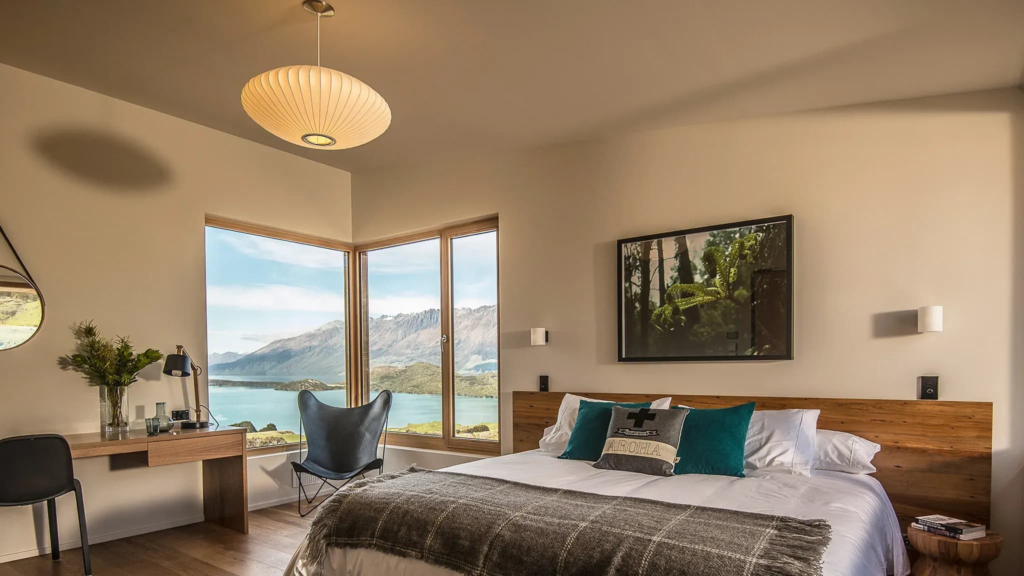 Timber bedroom with large windows showcasing mountain and lake views, a bed with white linens, and a desk area.