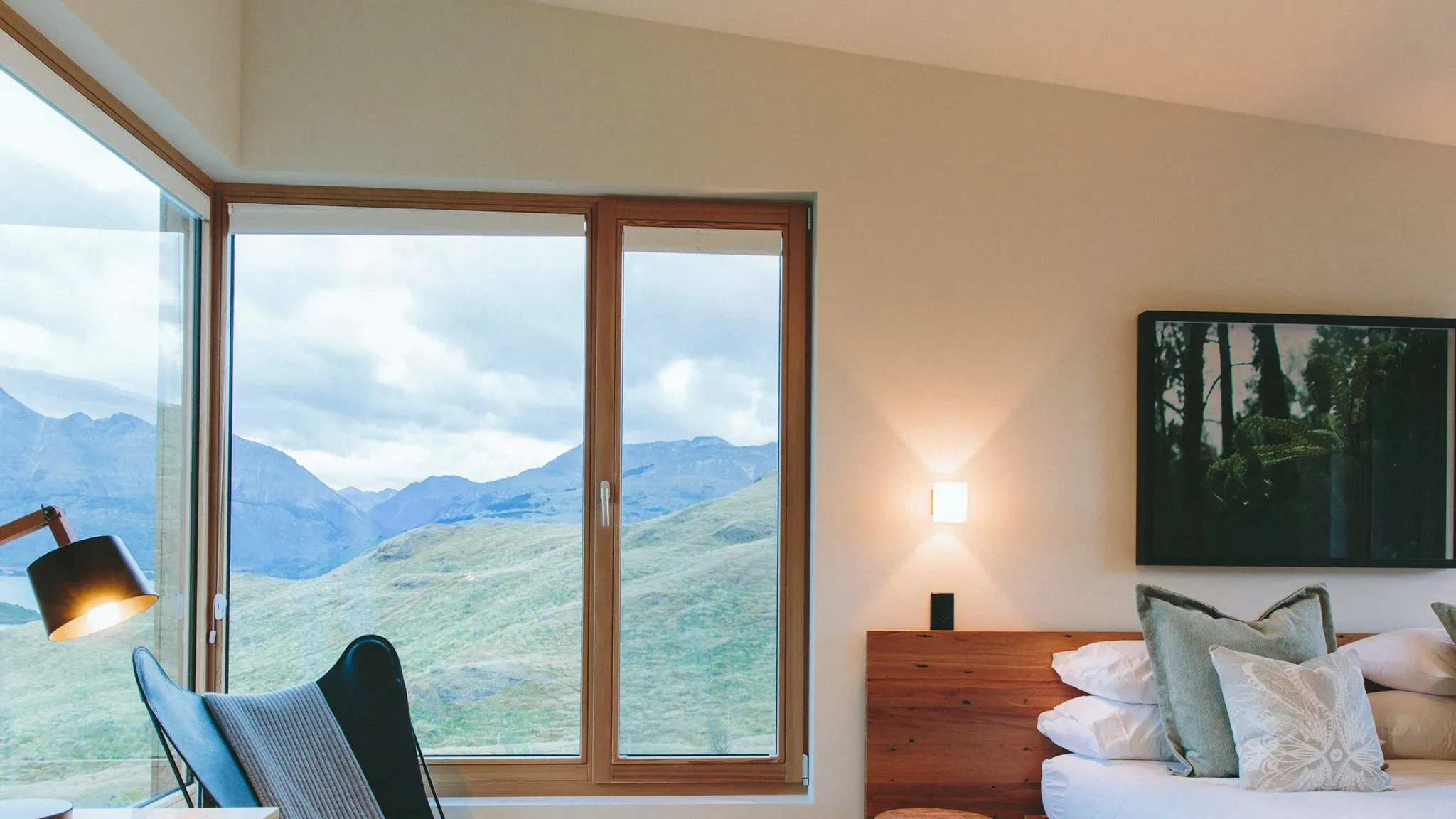 Timber-framed guest room with large windows showcasing mountain views, a bed with white and green pillows, and a wooden bedside table.