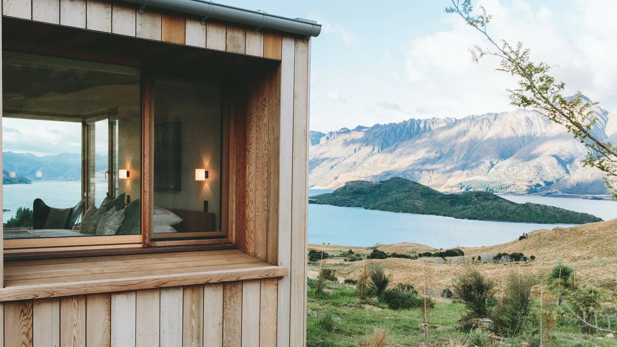 Timber structure with large window revealing a view of mountains and a lake, featuring soft furnishings inside.
