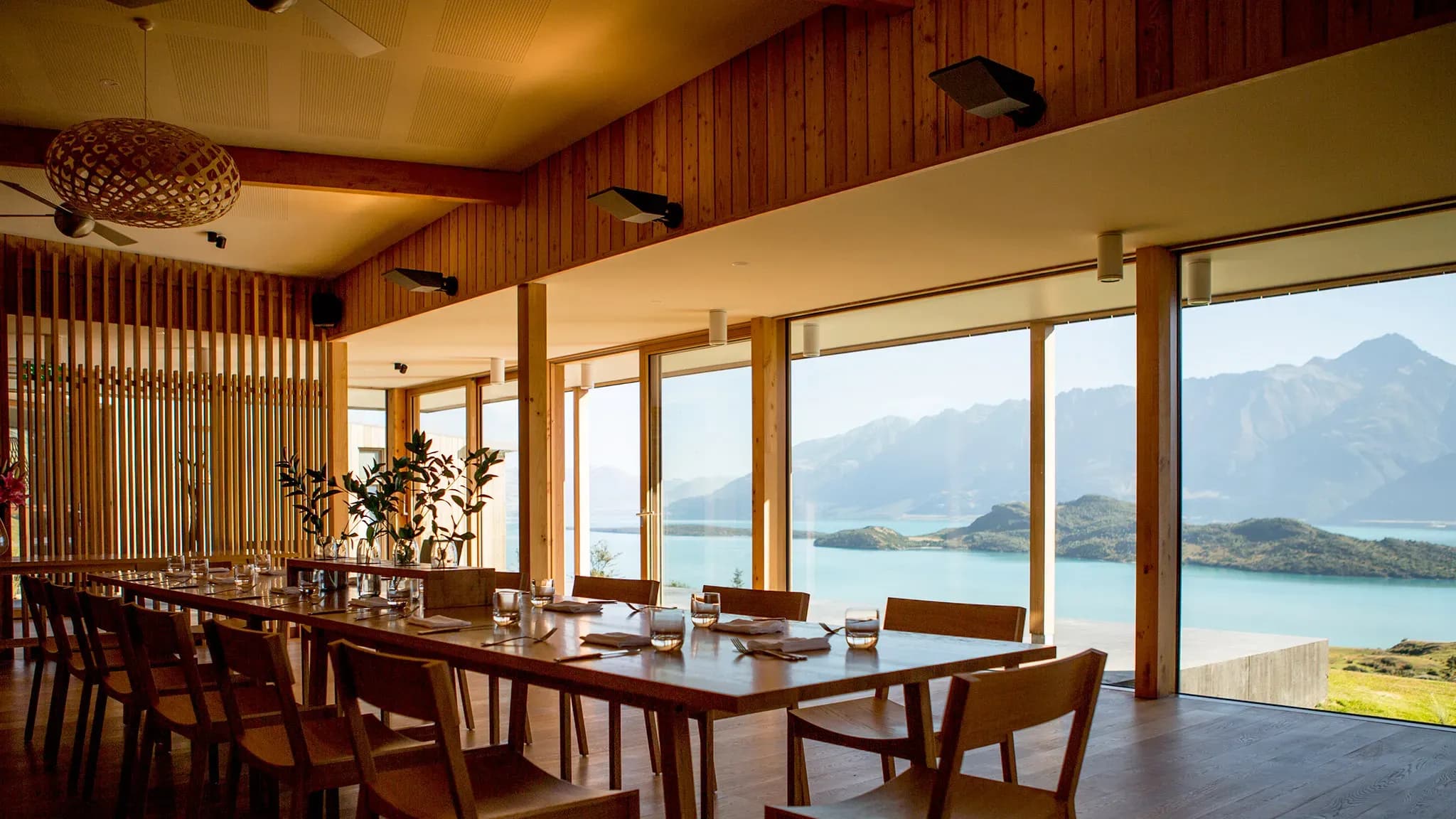Dining area with wooden table, chairs, and large windows showcasing views of mountains and water.