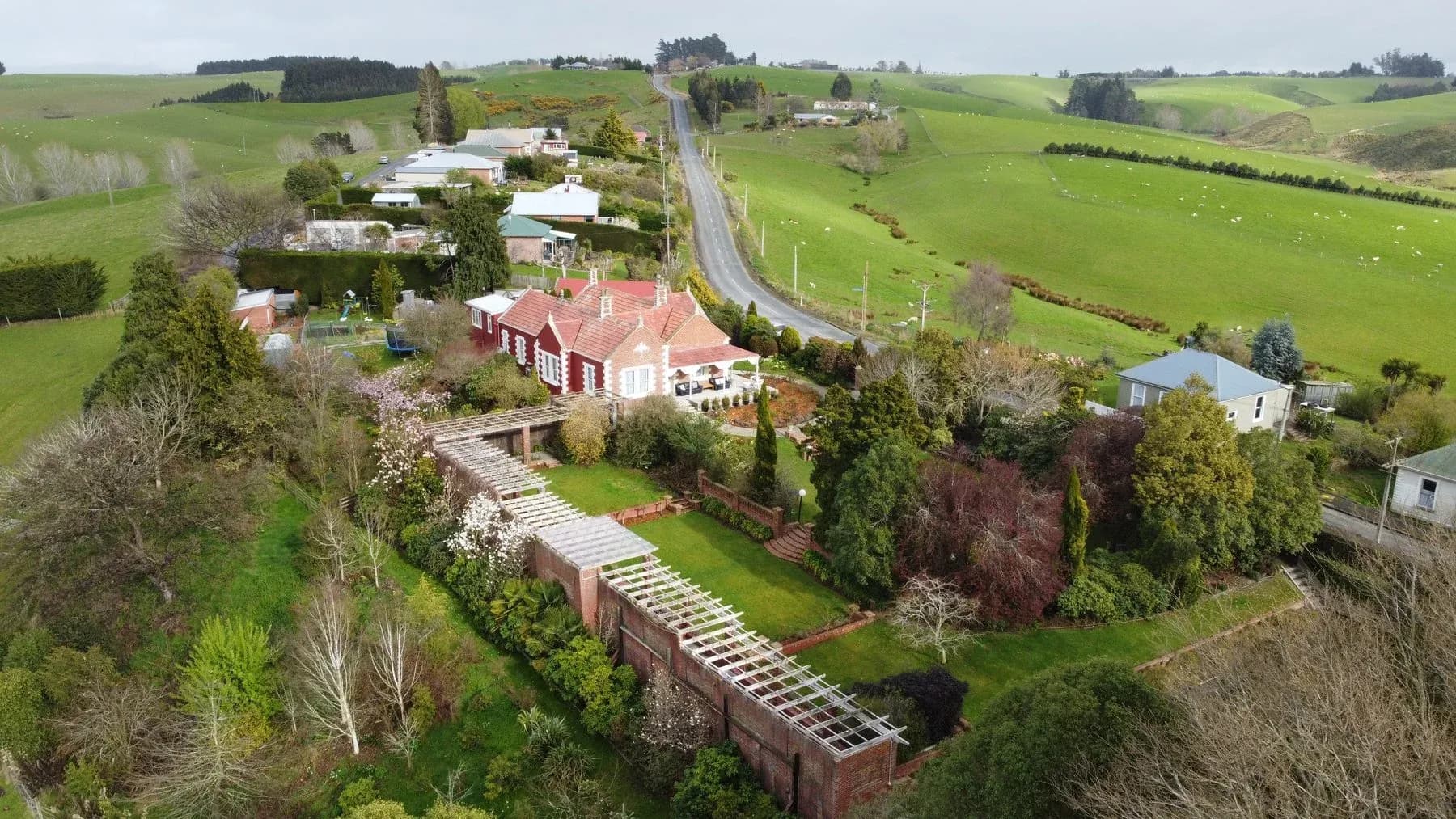 Aerial view of The Fern & Thistle villa surrounded by gardens, with green hills and a road in the background.