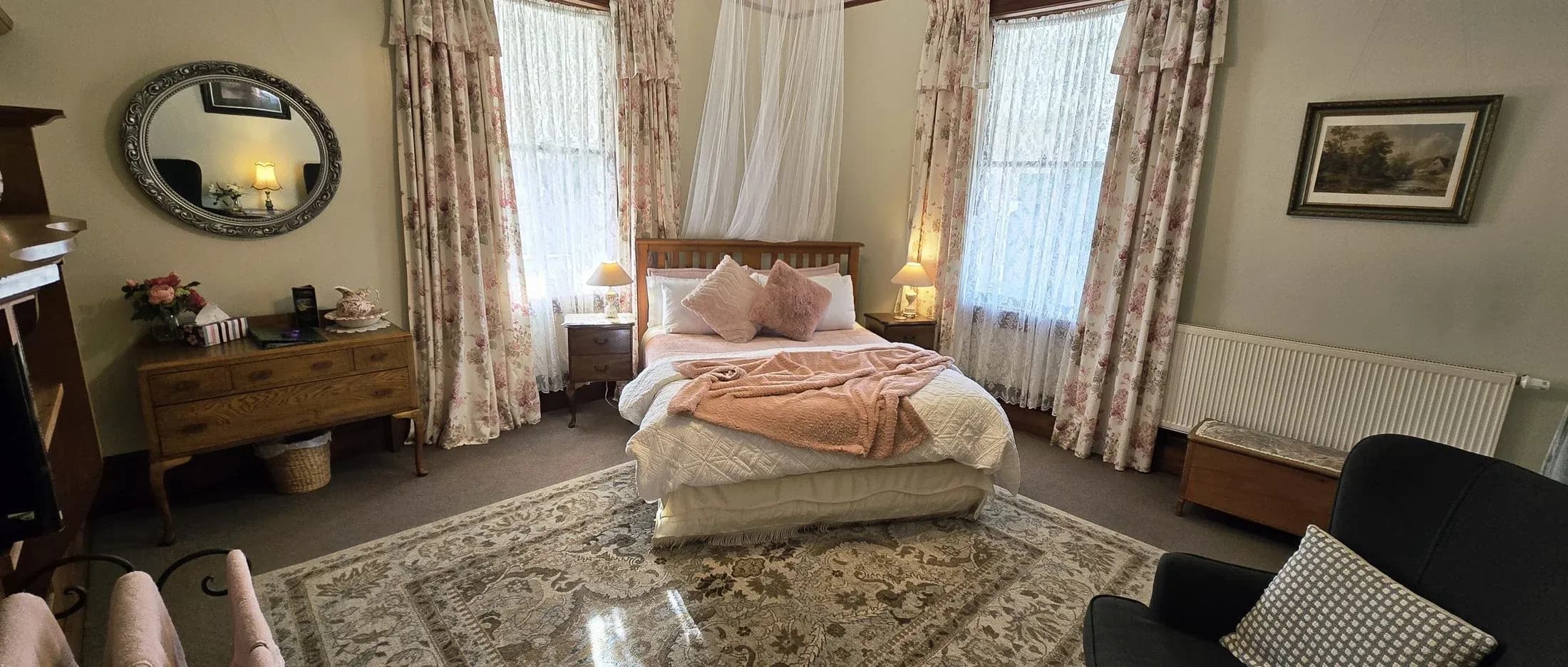 Timber-framed bedroom with floral curtains, a quilted bedspread, bedside lamps, and a decorative mirror reflecting soft light.