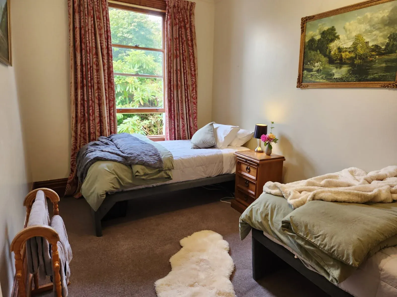 Two single beds with layered bedding and a bedside table in a room featuring a large window and floral curtains.