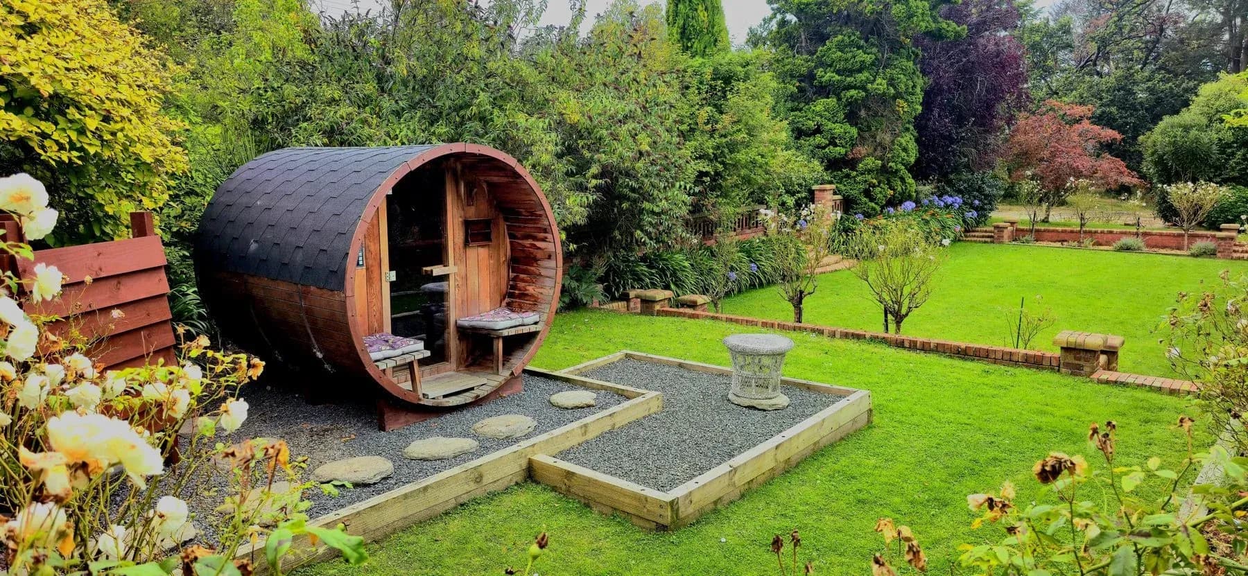 Barrel sauna situated in a garden with gravel paths, stone slabs, and surrounding greenery at The Fern & Thistle.