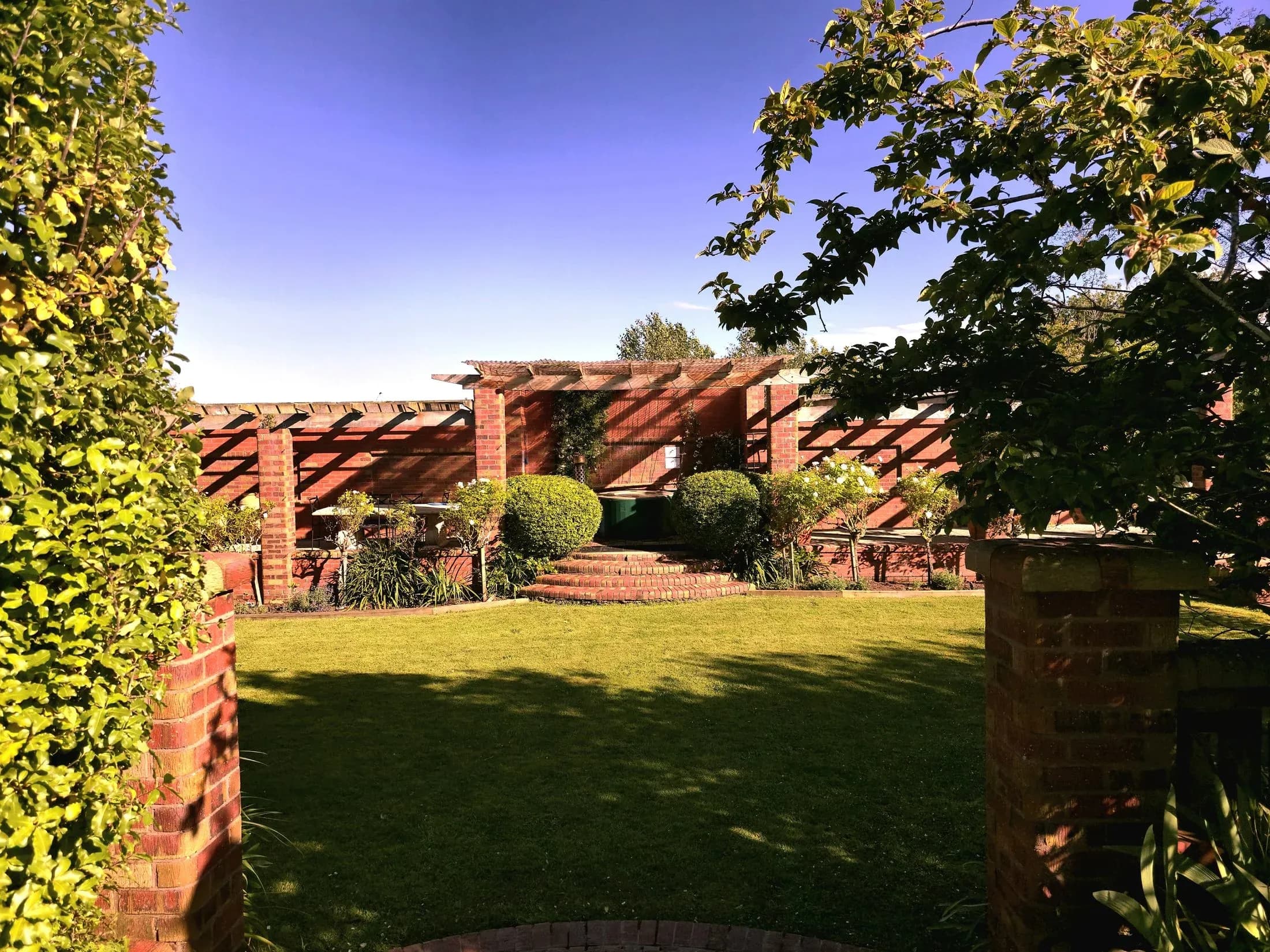 Garden pathway framed by brick walls and greenery, leading to a landscaped area with shrubs and a pergola.