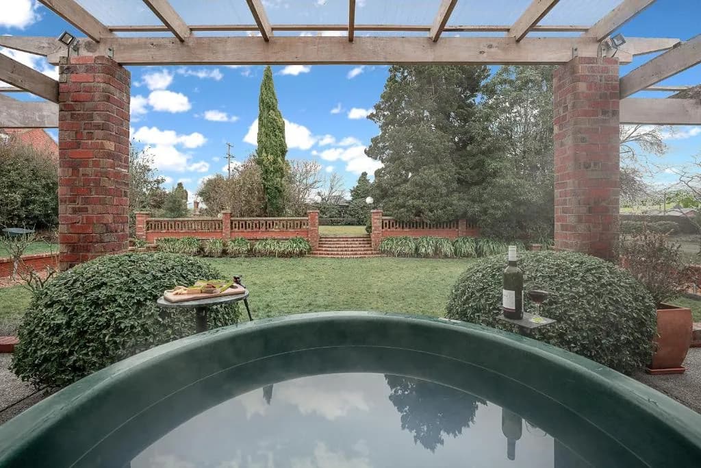 Barrel hot tub with a wine bottle and glass beside a wooden tray, overlooking a garden with brick pathways and greenery.