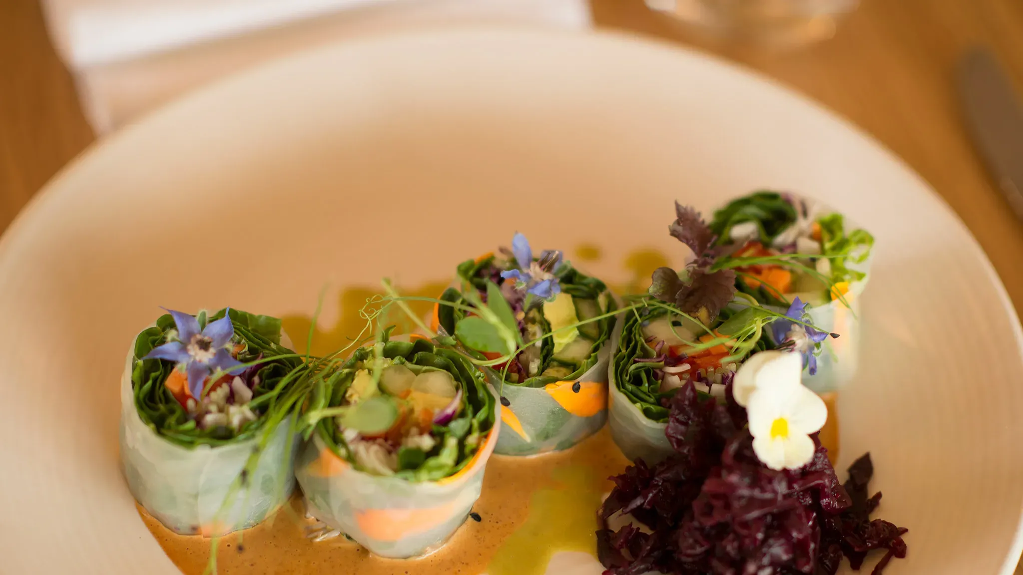 Fresh vegetable rolls with greens, edible flowers, and a side of shredded beetroot on a cream plate.