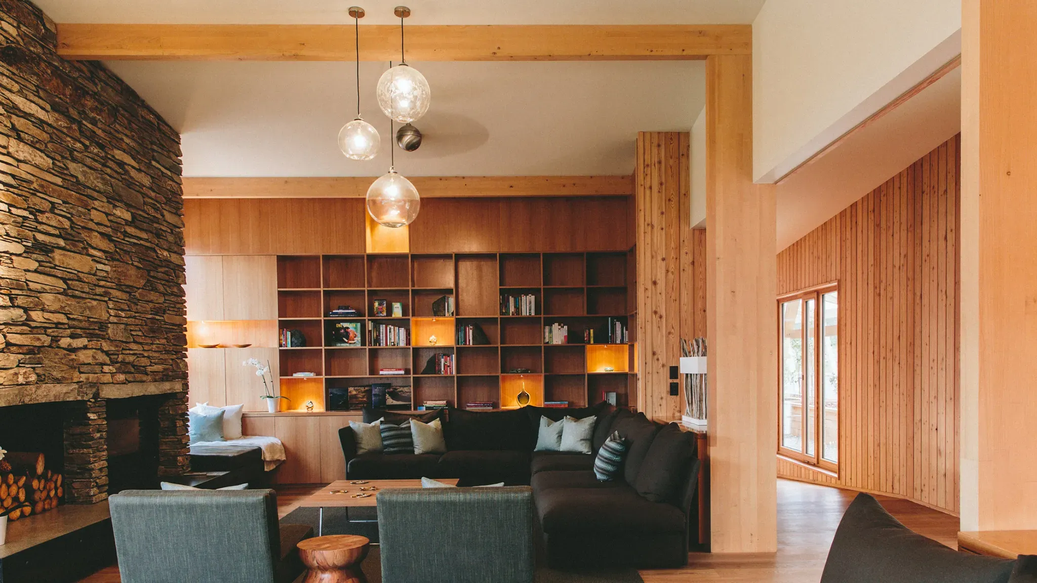 Seating area with a dark sectional sofa, wooden coffee table, and stone fireplace, surrounded by wooden bookshelves and large windows.