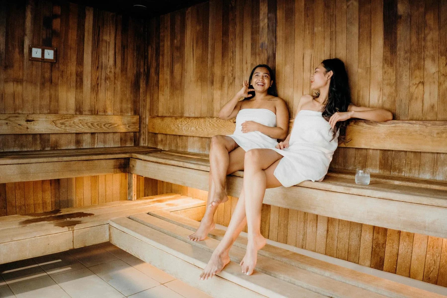 Two guests seated on wooden bench in a sauna, wearing white towels, with wooden walls and warm lighting.