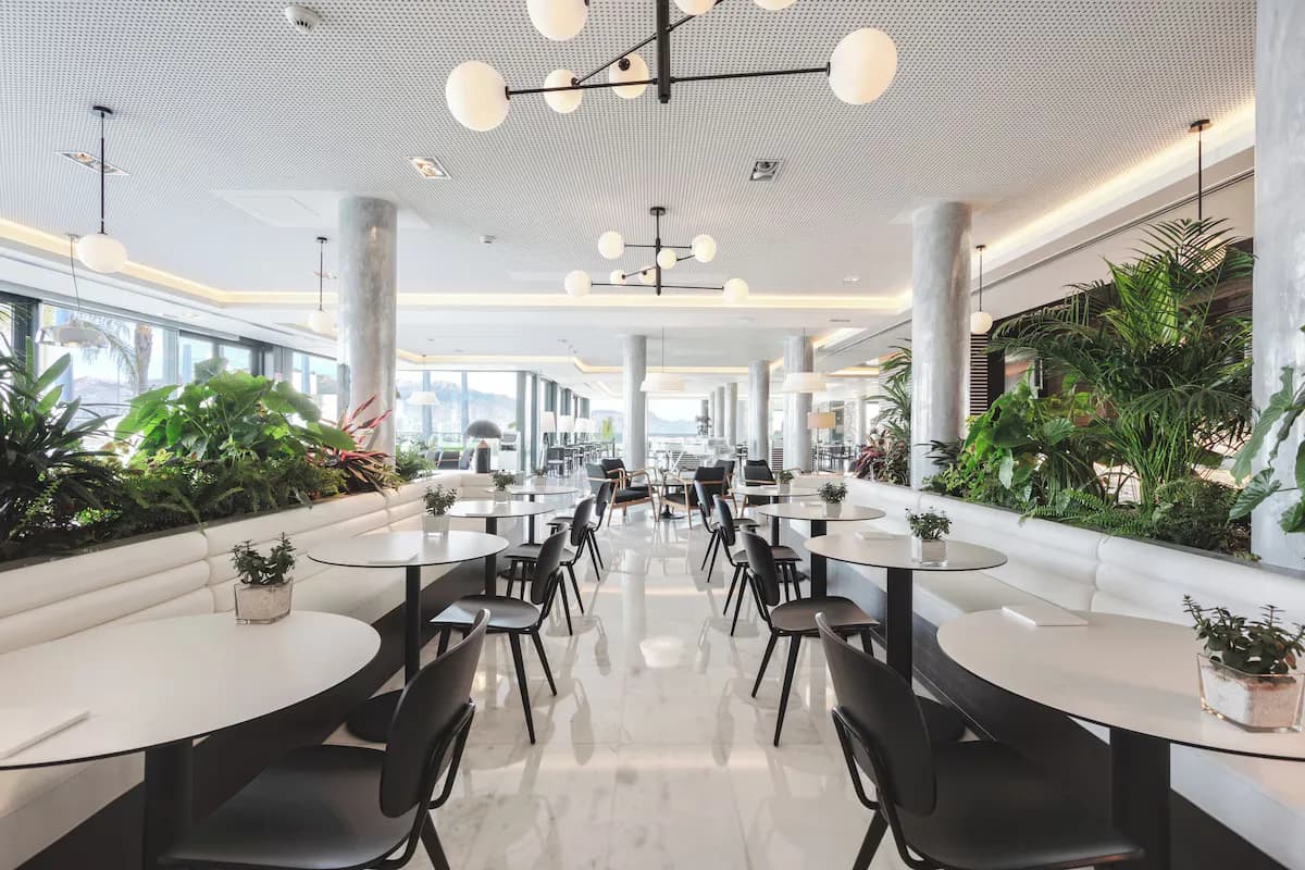 Dining area with white marble floors, round tables, black chairs, and greenery in planters under large windows.