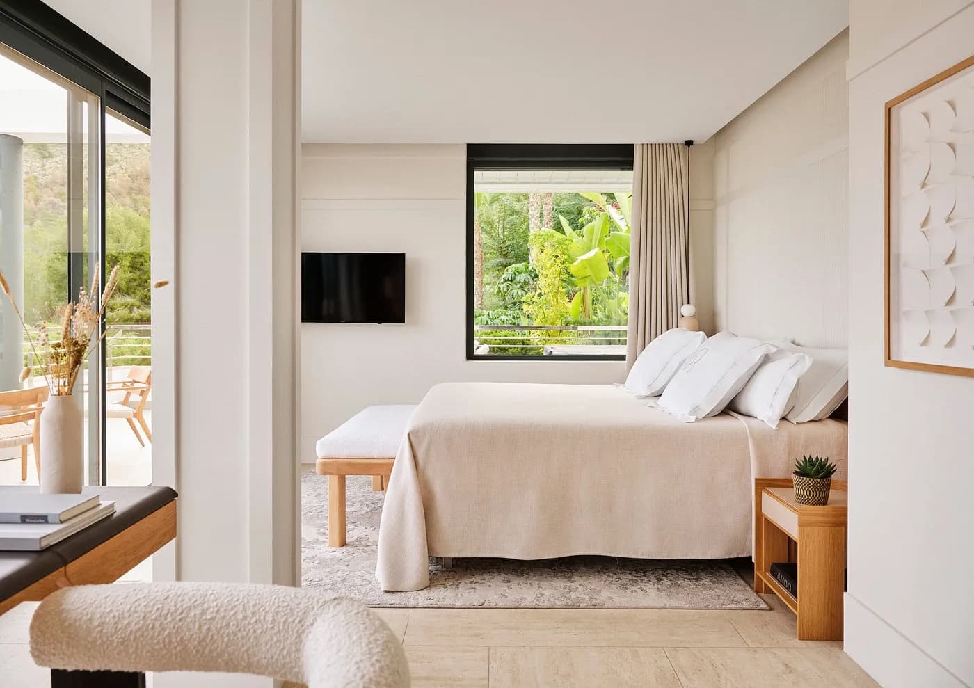 Bright bedroom with a made bed, neutral bedding, a television, and large windows overlooking greenery.