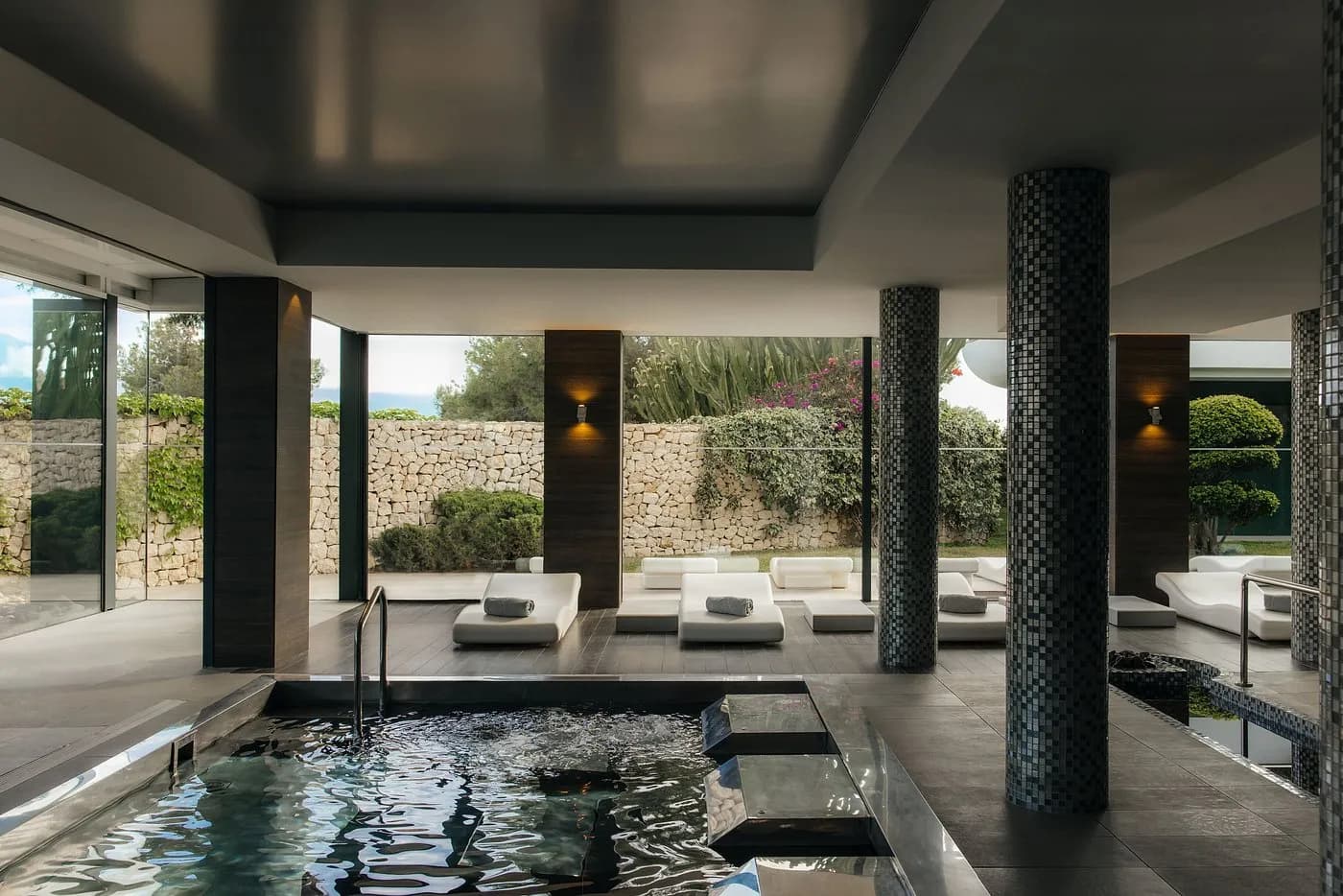 Indoor hydrotherapy pool surrounded by stone walls, with lounge chairs and large windows overlooking greenery.