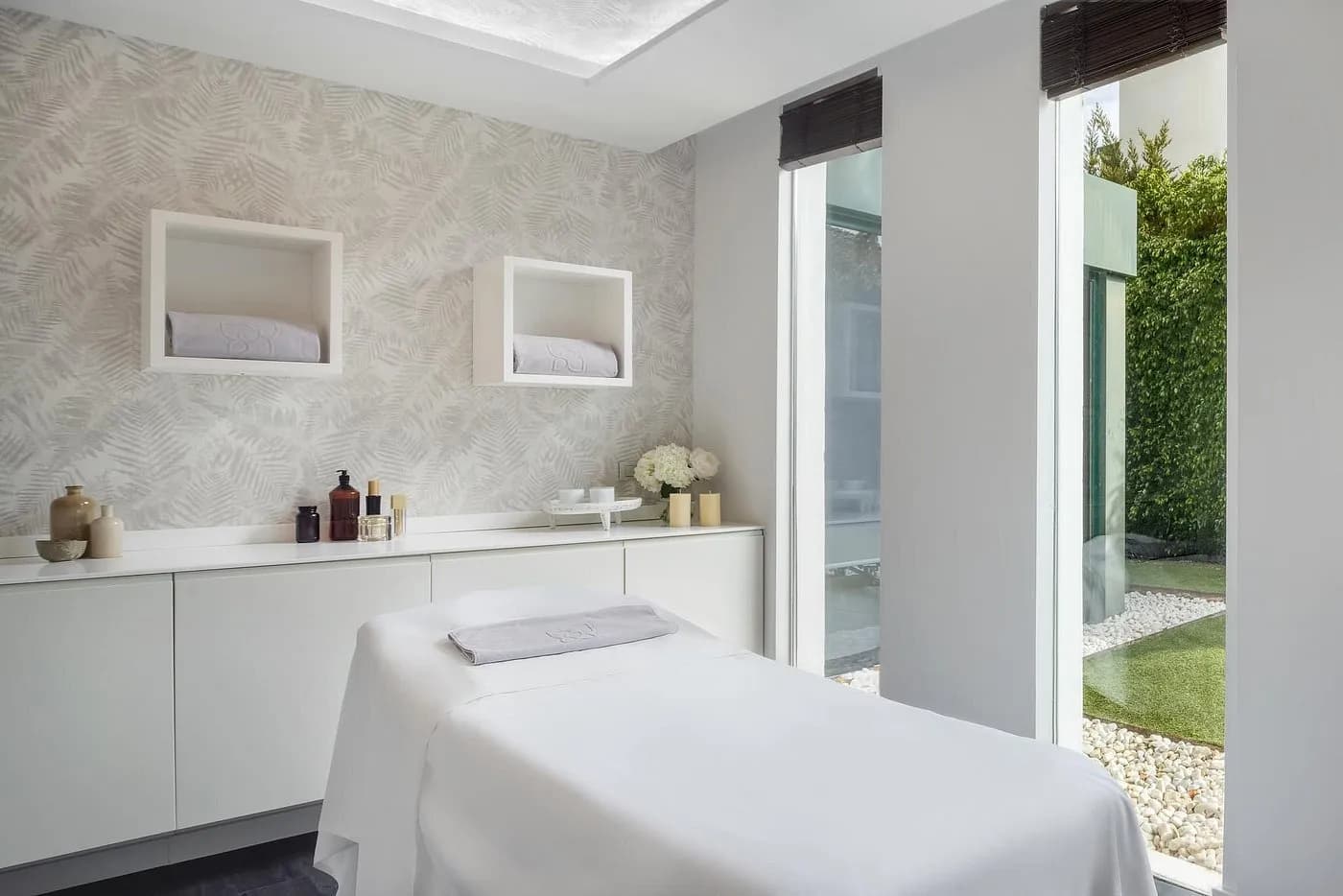 Spa treatment room with a massage table, light-colored cabinetry, decorative bottles, and large windows overlooking a garden.