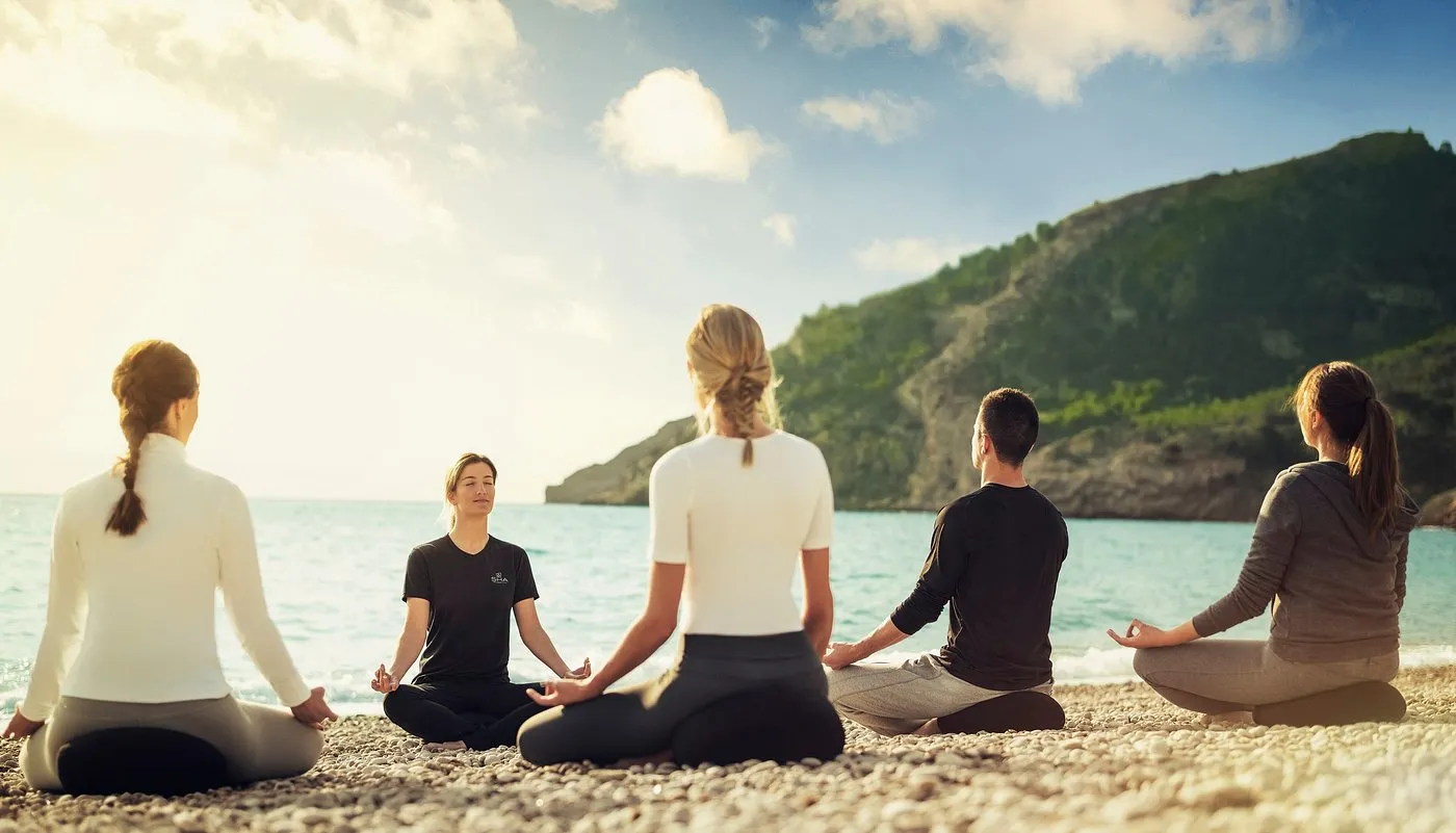Guests meditating on a pebble beach with the sea and a hillside in the background during sunset.