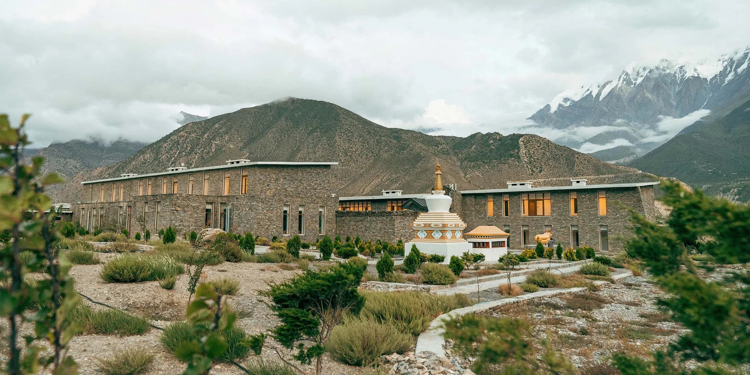 Stone building with large windows, surrounded by sparse vegetation and mountains under a cloudy sky.
