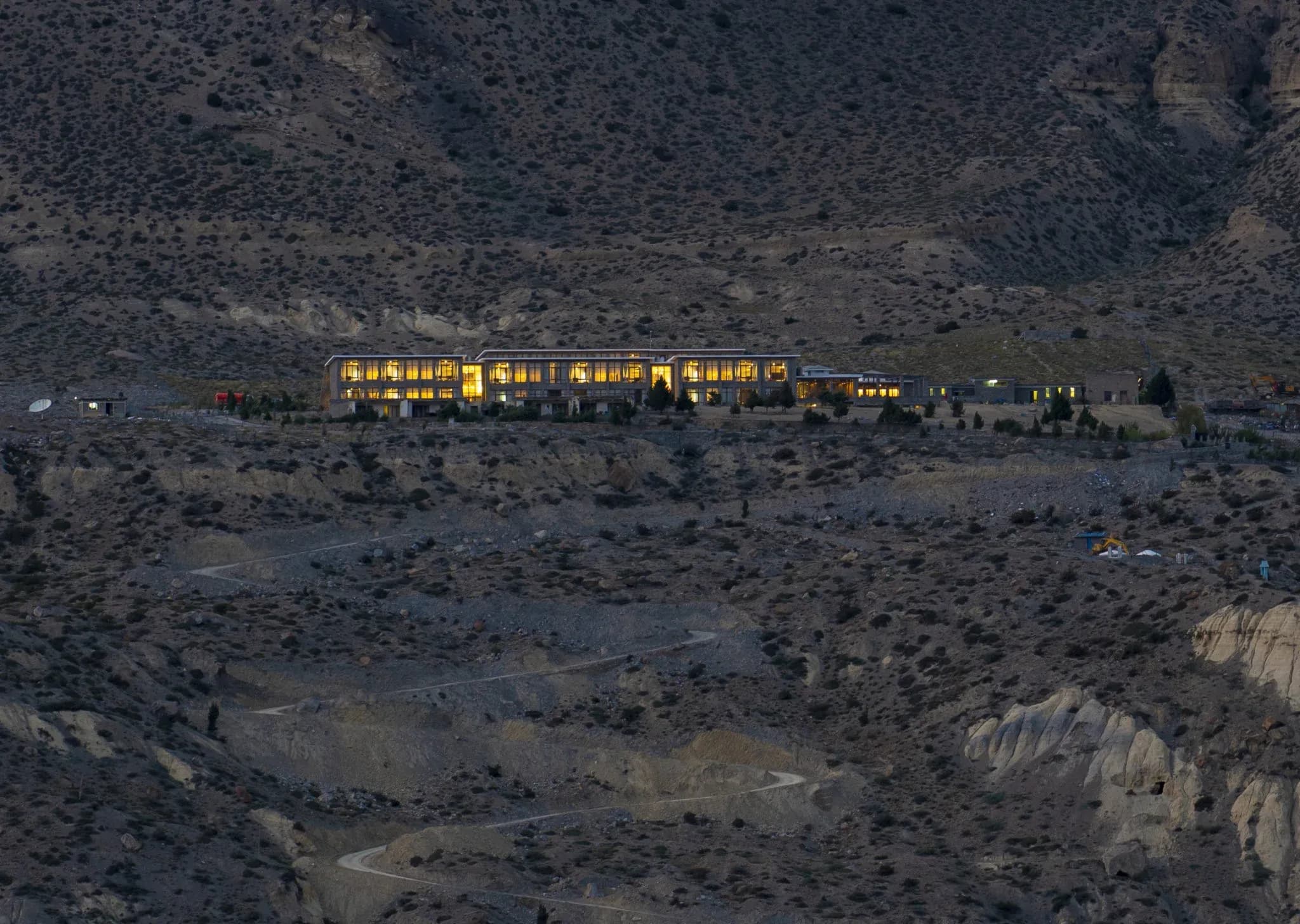 Lodge exterior illuminated at dusk, set against a rugged mountainous landscape with winding paths and sparse vegetation.
