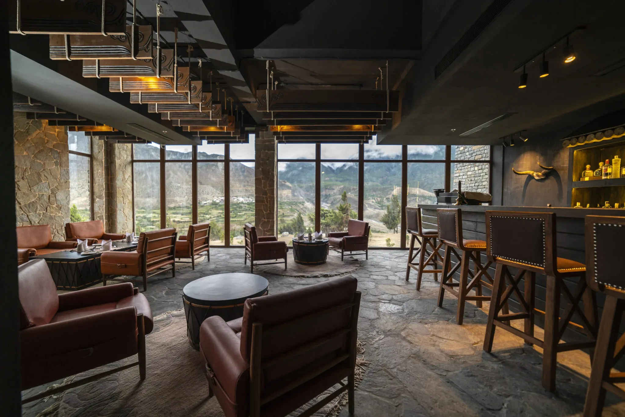 Lounge area with stone walls, leather seating, and a bar featuring backlit shelves, overlooking mountainous landscapes through large windows.