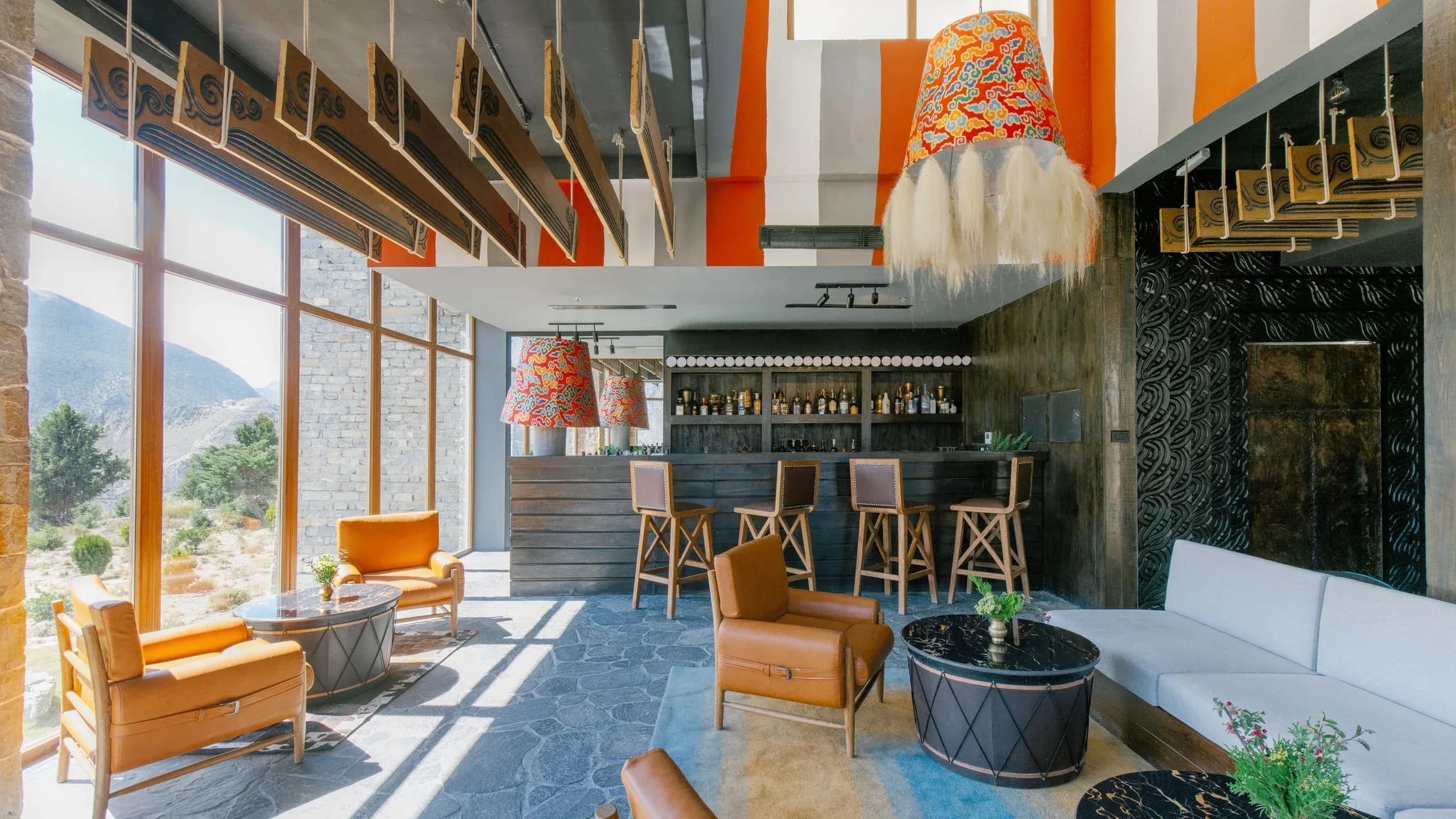 Lounge area with large windows, orange-accented furniture, patterned light fixtures, and a wooden bar with bottles.