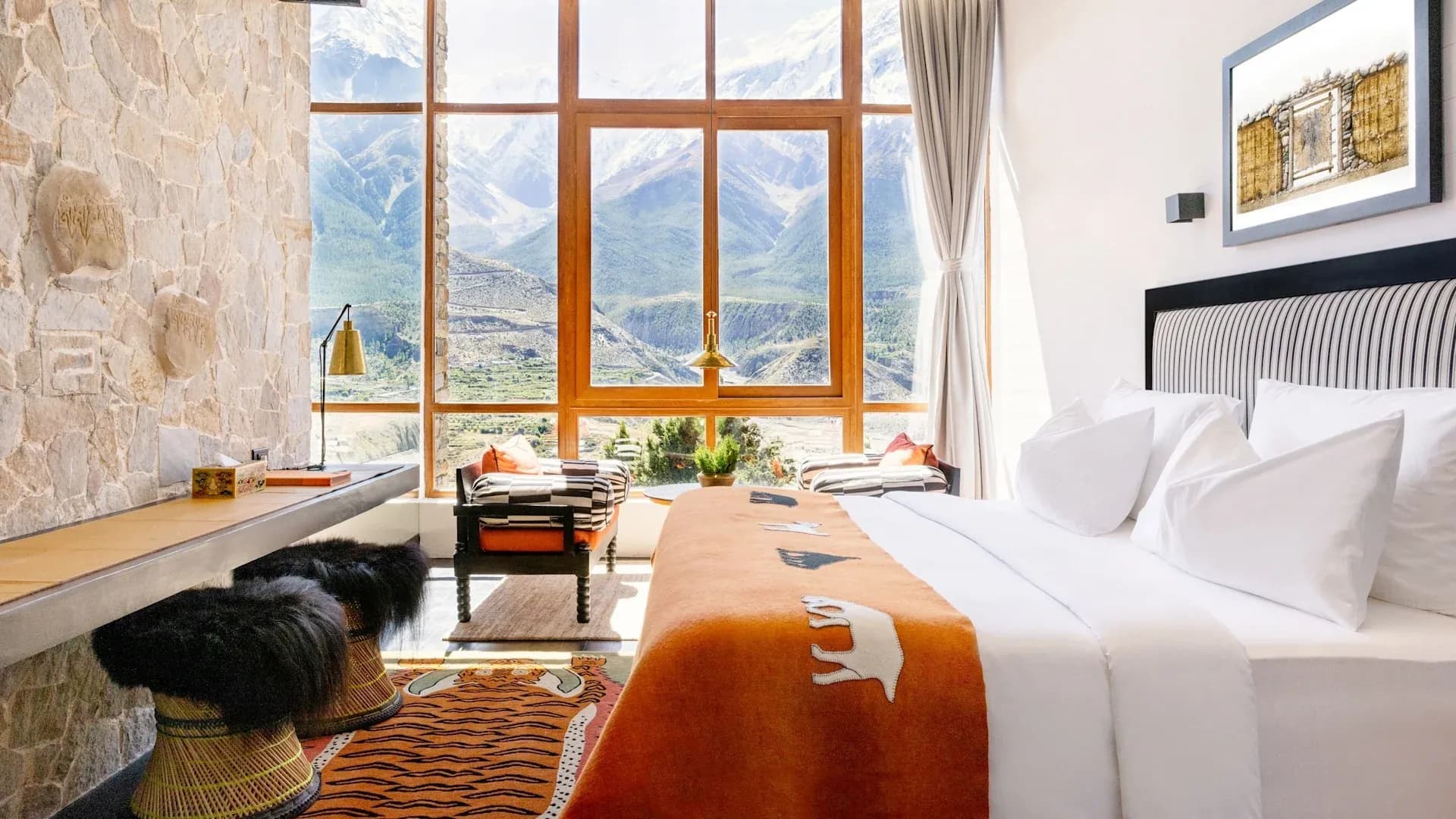 Timber-framed bedroom with a stone accent wall, white bedding, orange throw, and large windows showcasing mountain views.