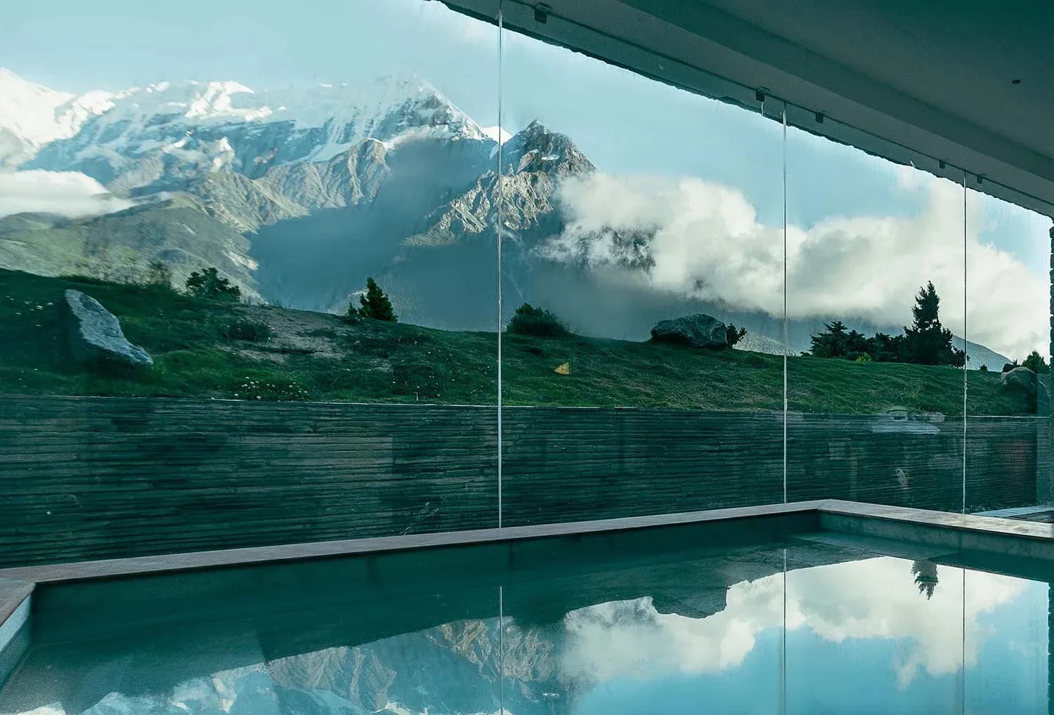 Indoor pool area with glass walls reflecting snow-capped mountains and green hillside in the background.