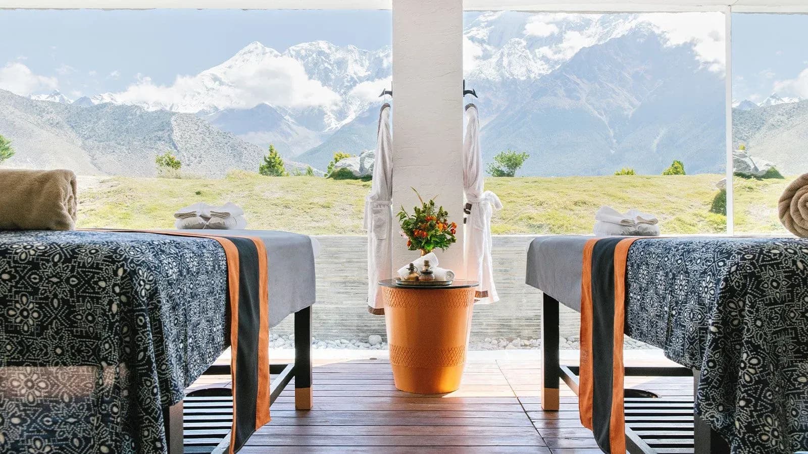 Two treatment tables with patterned covers and orange accents, a potted flower arrangement, and mountain views through large windows.