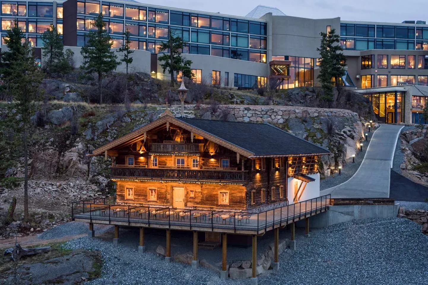Timber building with a large deck, surrounded by gravel and trees, with a modern resort structure above.