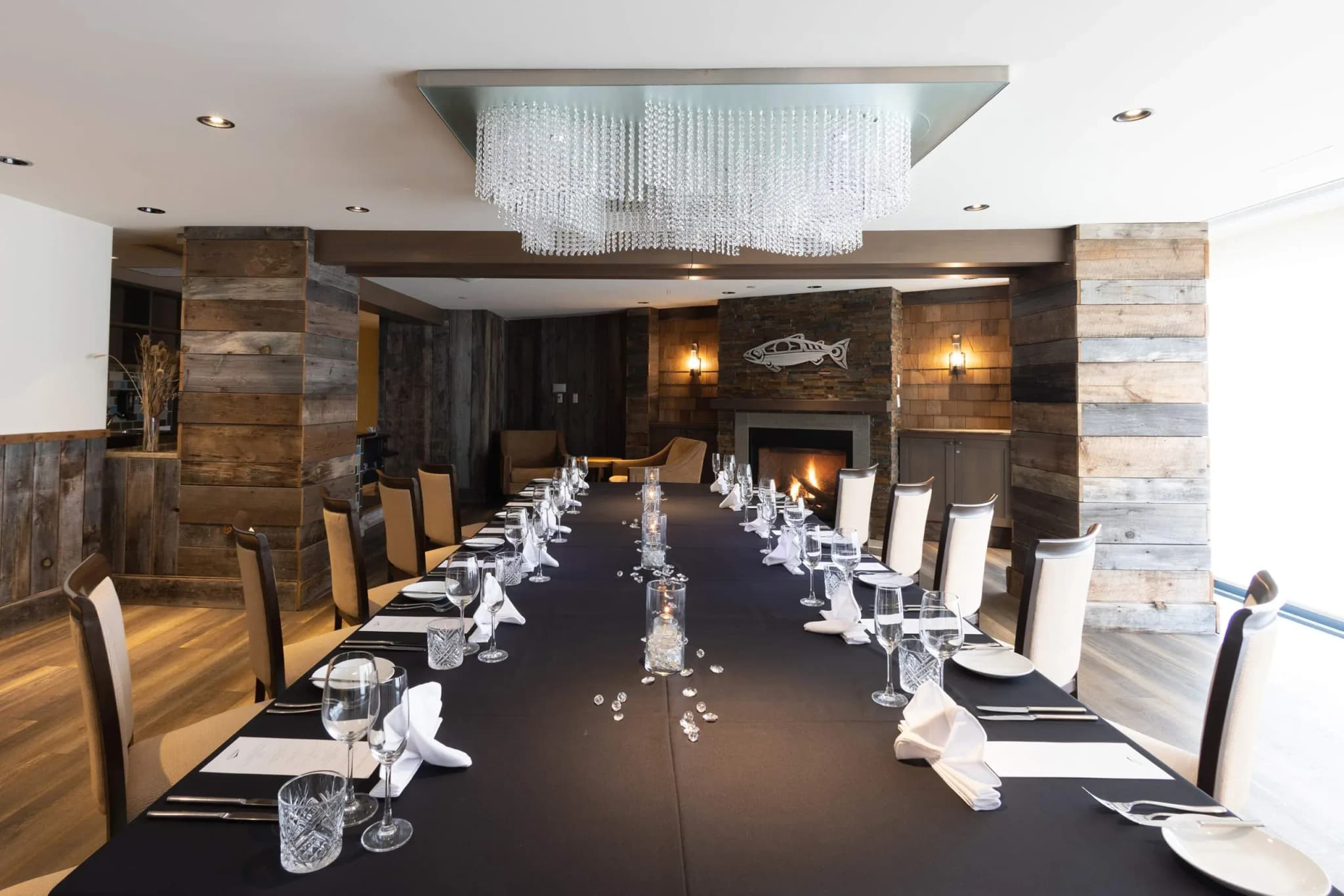 Long dining table set with black tablecloth, white napkins, and crystal glasses, under a chandelier, with a fireplace in the background.