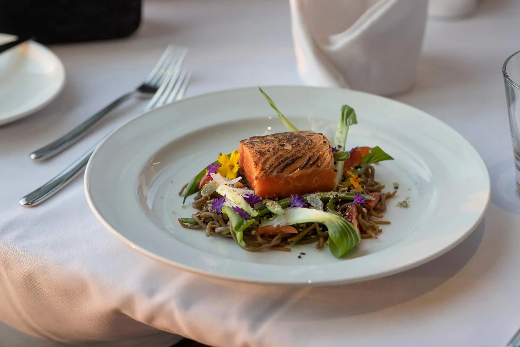 Grilled salmon fillet atop soba noodles, garnished with green vegetables and edible flowers on a white plate.