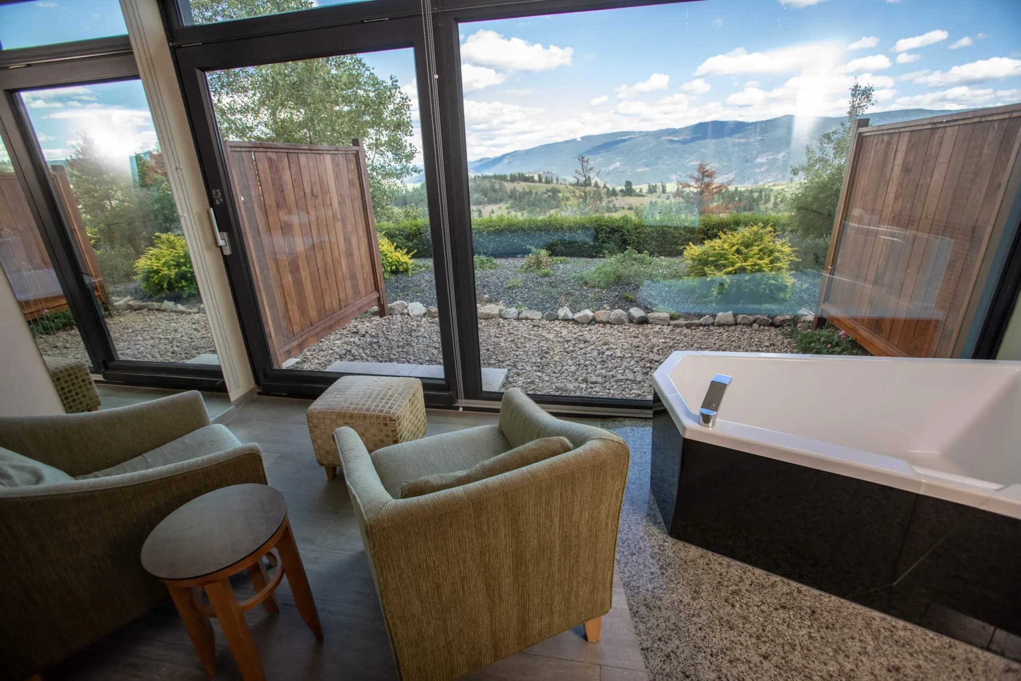 Seating area with two green armchairs and a wooden table beside a window overlooking landscaped grounds and mountains.
