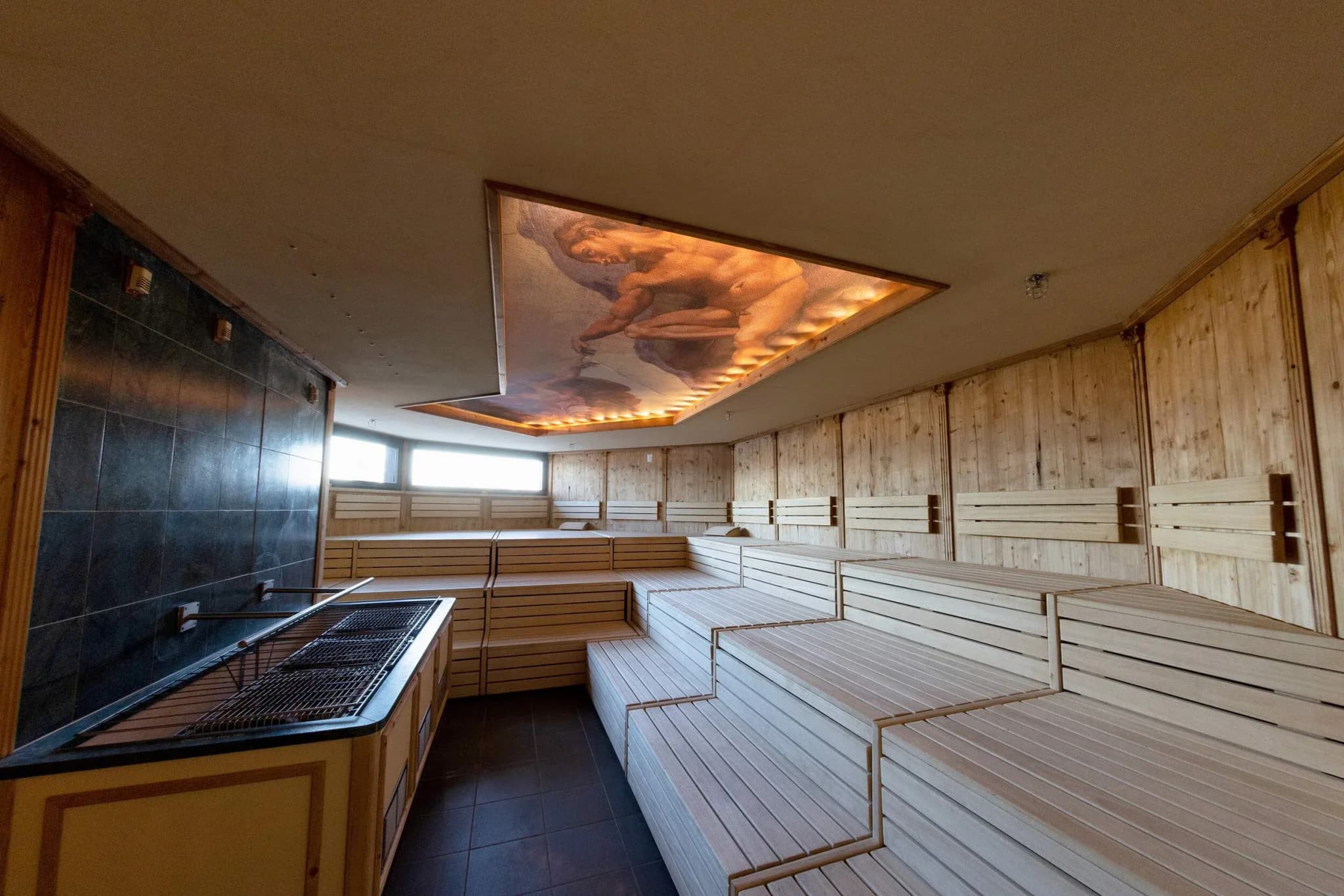 Timber sauna interior with tiered wooden benches, a stone wall, and a decorative ceiling light featuring a mural.