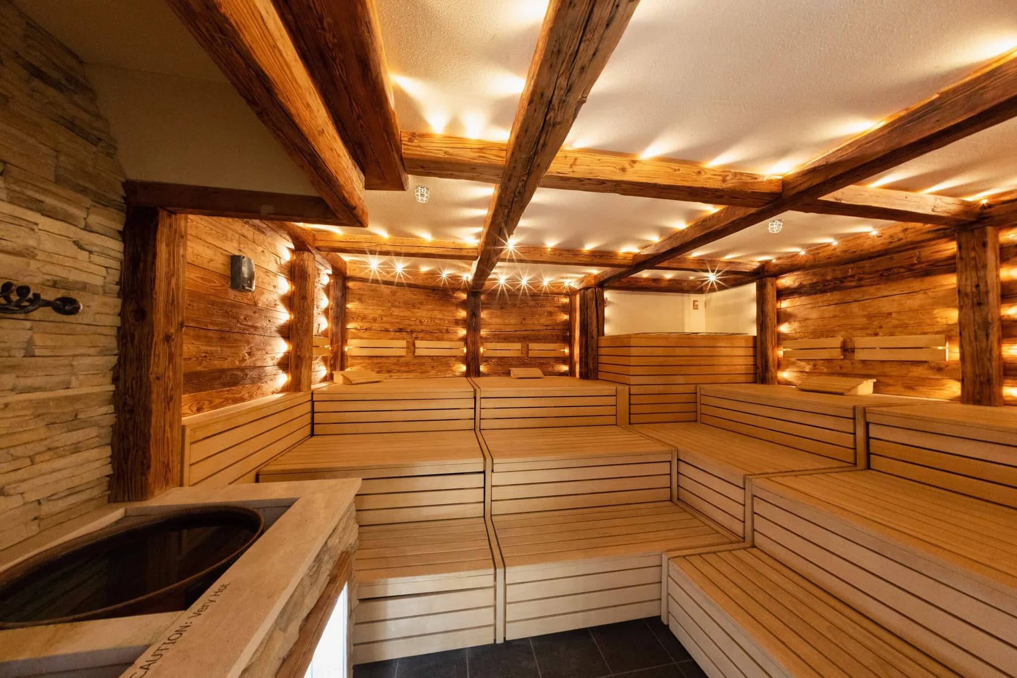 Timber sauna interior with tiered wooden seating, stone wall, ambient lighting, and a shallow plunge pool.