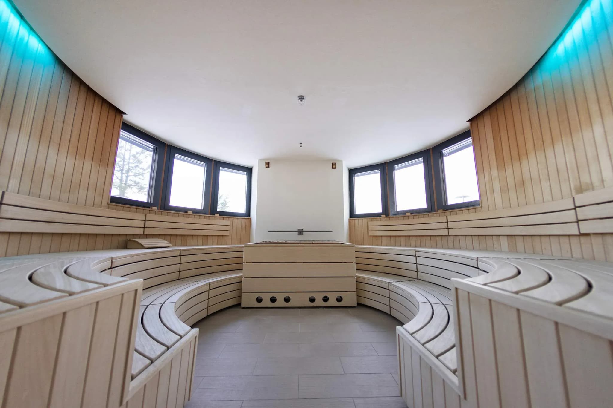 Timber sauna interior featuring curved bench seating, large windows, and neutral tile flooring.