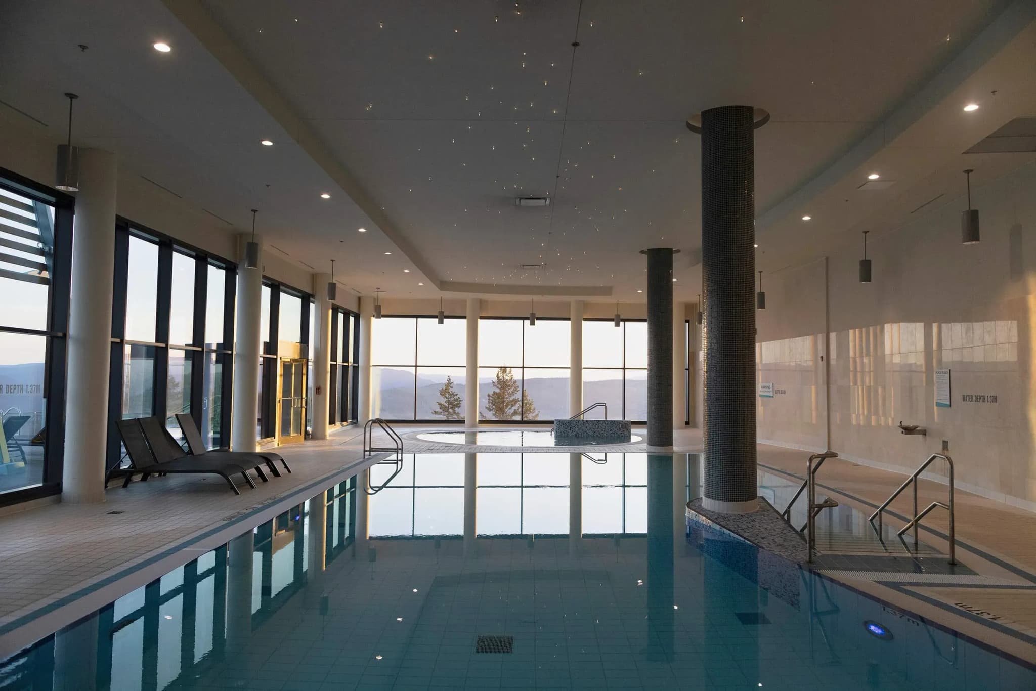 Indoor swimming pool area with large windows, lounge chairs, and a hot tub, overlooking distant hills.