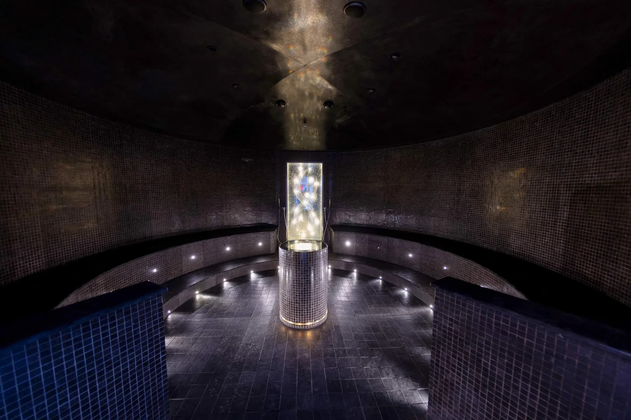 Steam room interior with curved seating, mosaic tiles, and ambient lighting surrounding a central illuminated feature.