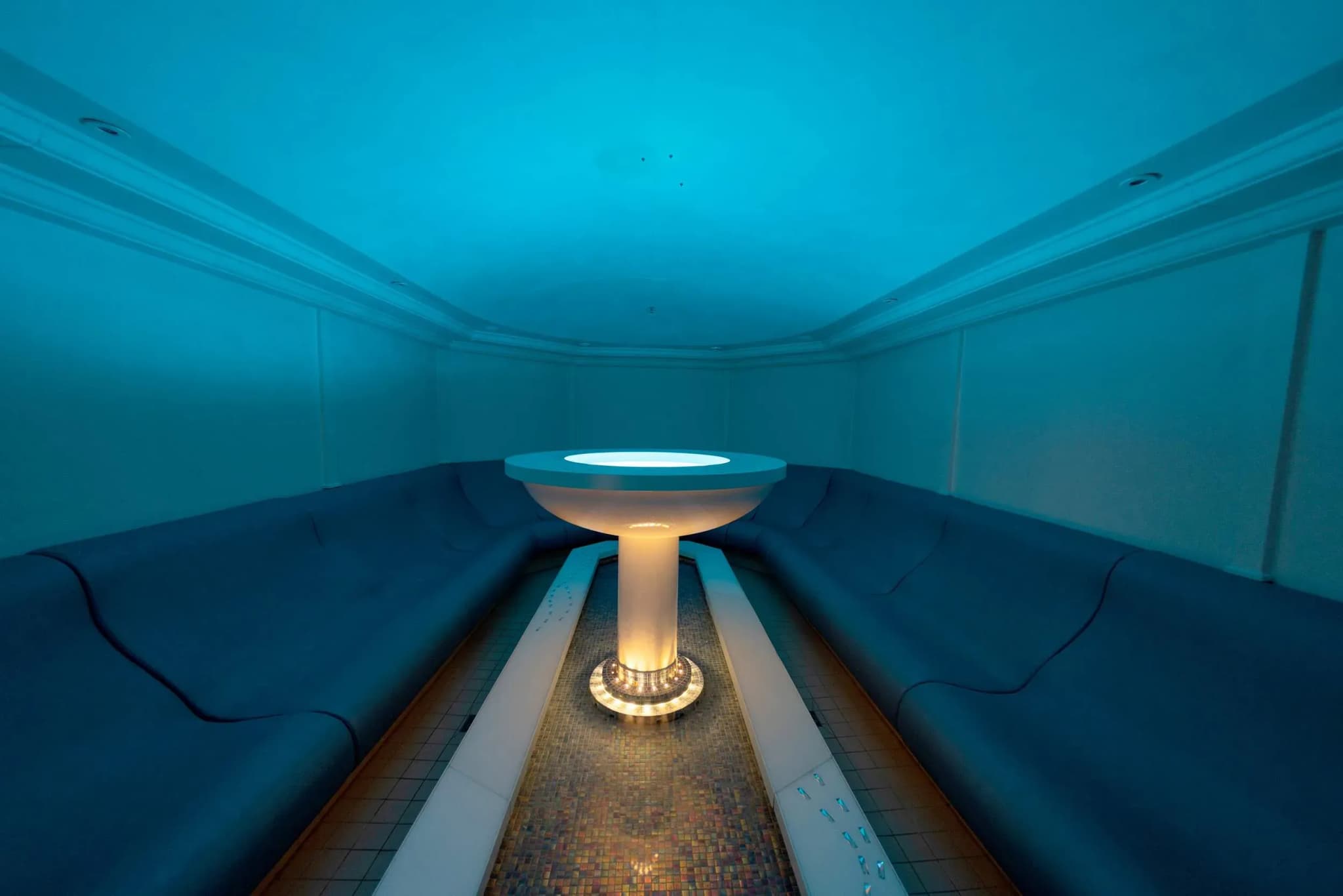 Indoor steam room featuring curved seating in blue tones, illuminated central basin, and mosaic tile flooring.