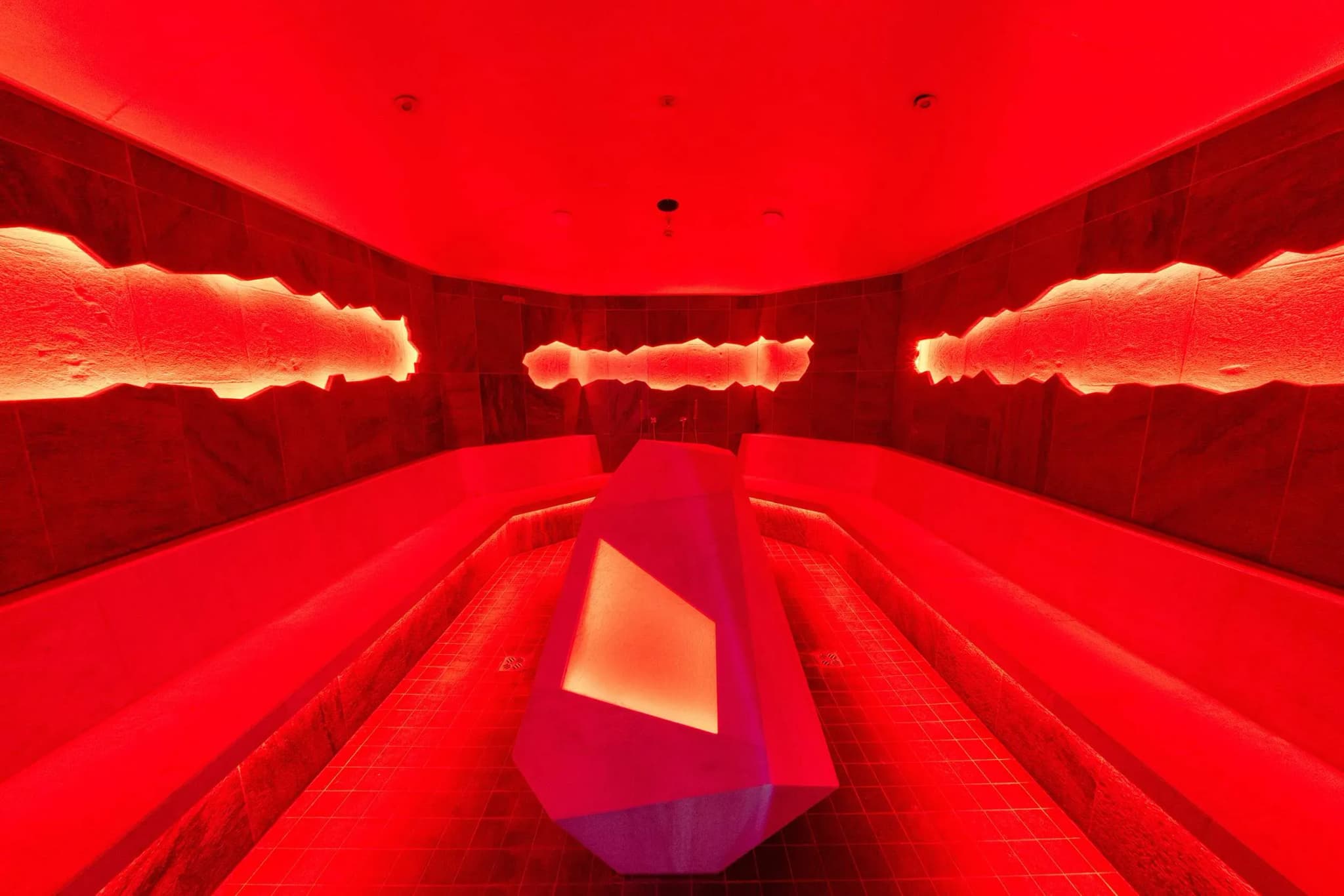 Interior of a steam room with red lighting, featuring curved benches and a central stone table.