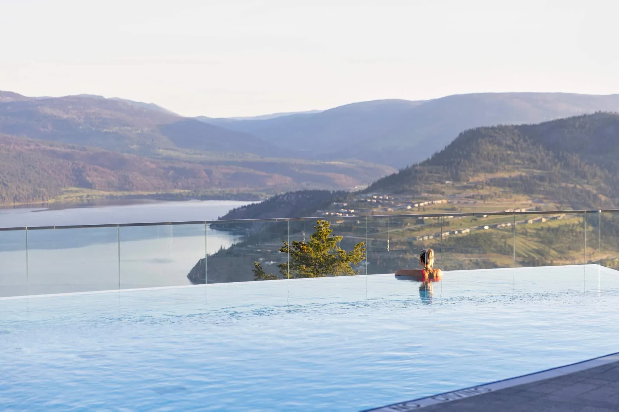 A guest relaxing in an infinity pool overlooking Okanagan Lake and surrounding mountains, with clear skies above.